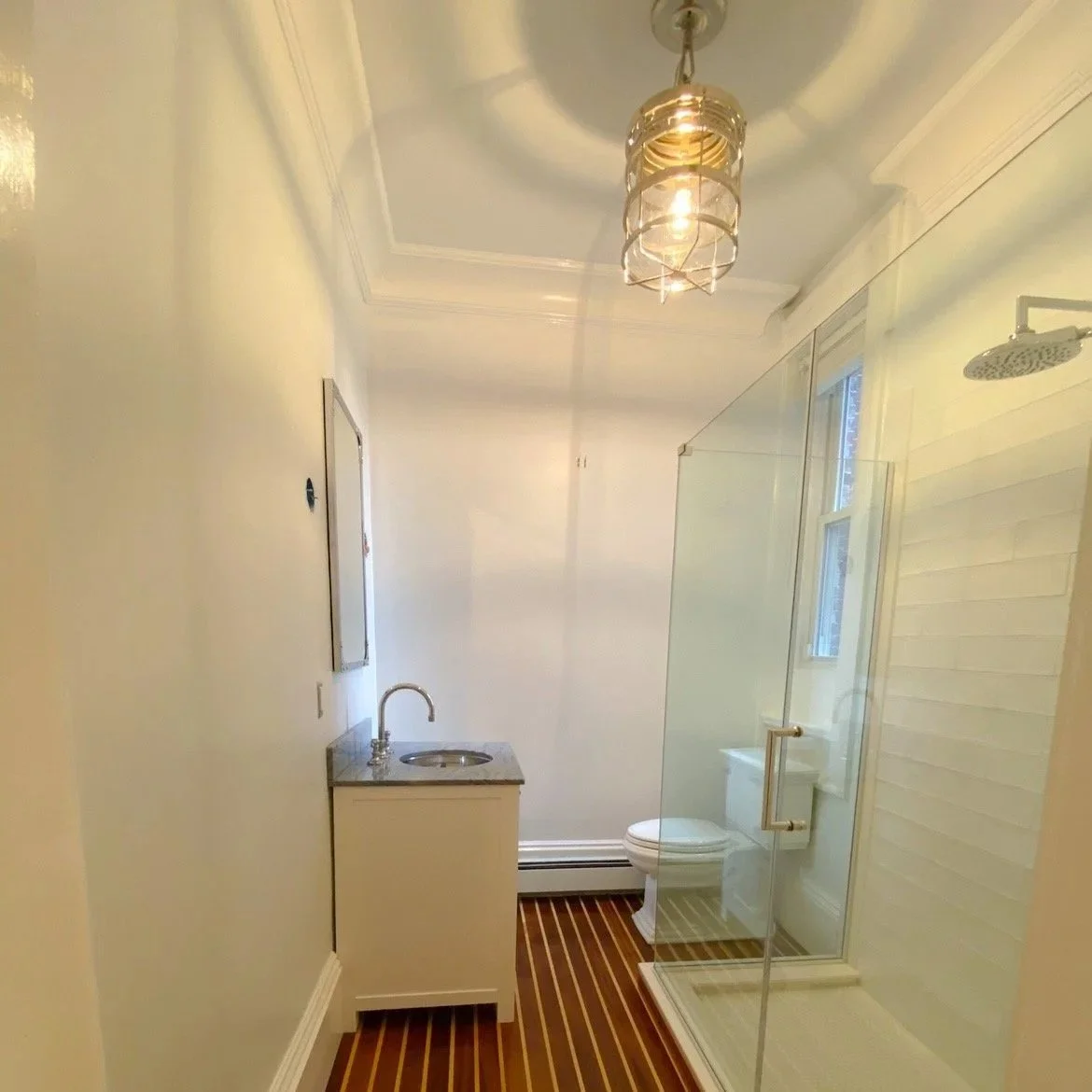 Bathroom with a shower, toilet, small sink with a granite top, a ceiling light fixture, and a window, with wood flooring. Renovated by Structure Pro in Massachusetts