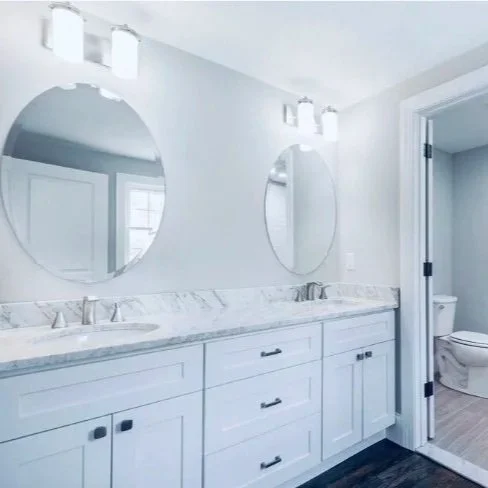 A modern bathroom with a double vanity, white cabinets, granite countertop, two oval mirrors, and a separate toilet area visible through an open door. Built Structure Pro in Massachusetts