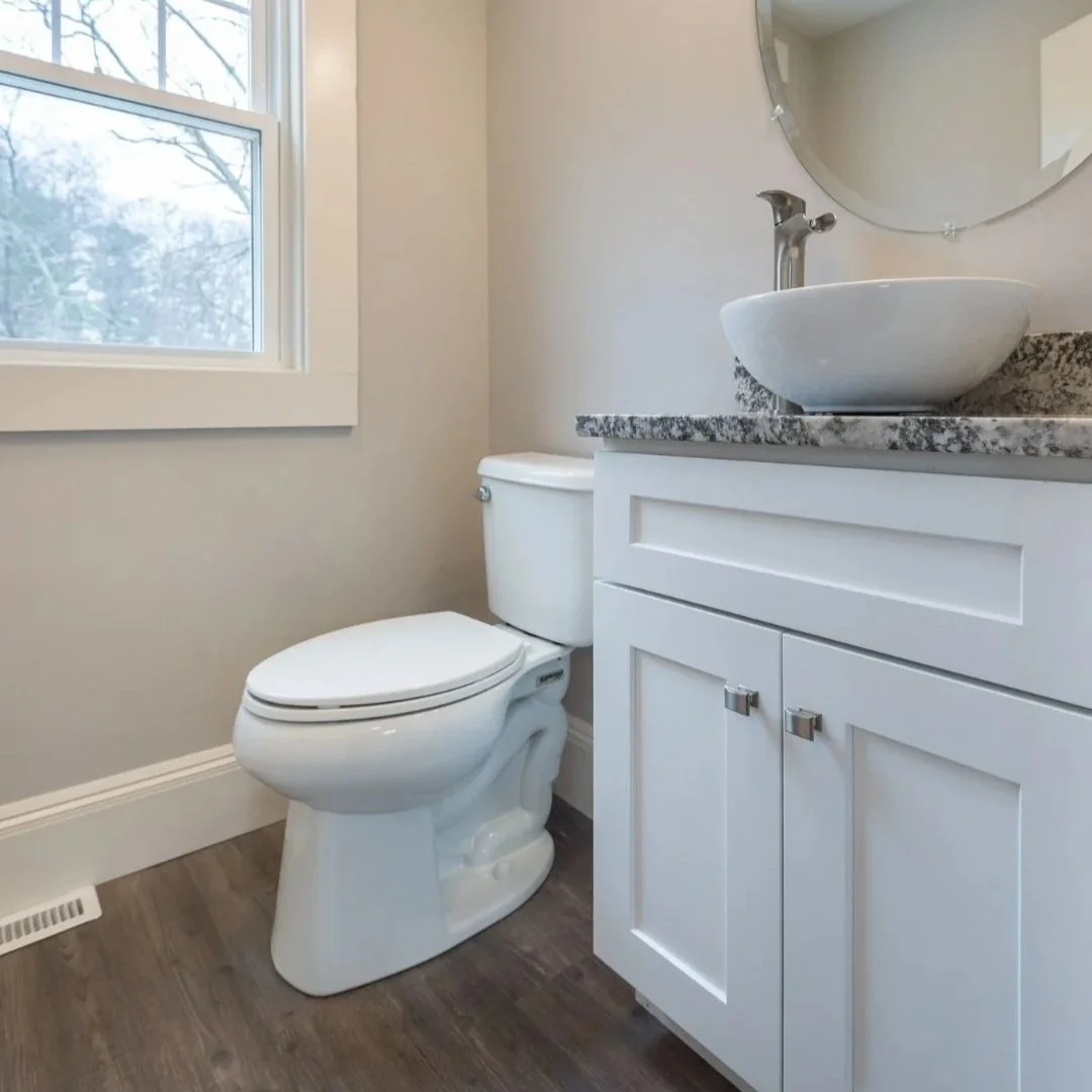 A small bathroom featuring a white toilet, a white vanity with a granite countertop, a vessel sink, and a round mirror above the sink. There is a window on the left and a wooden floor. Built Structure Pro in Massachusetts