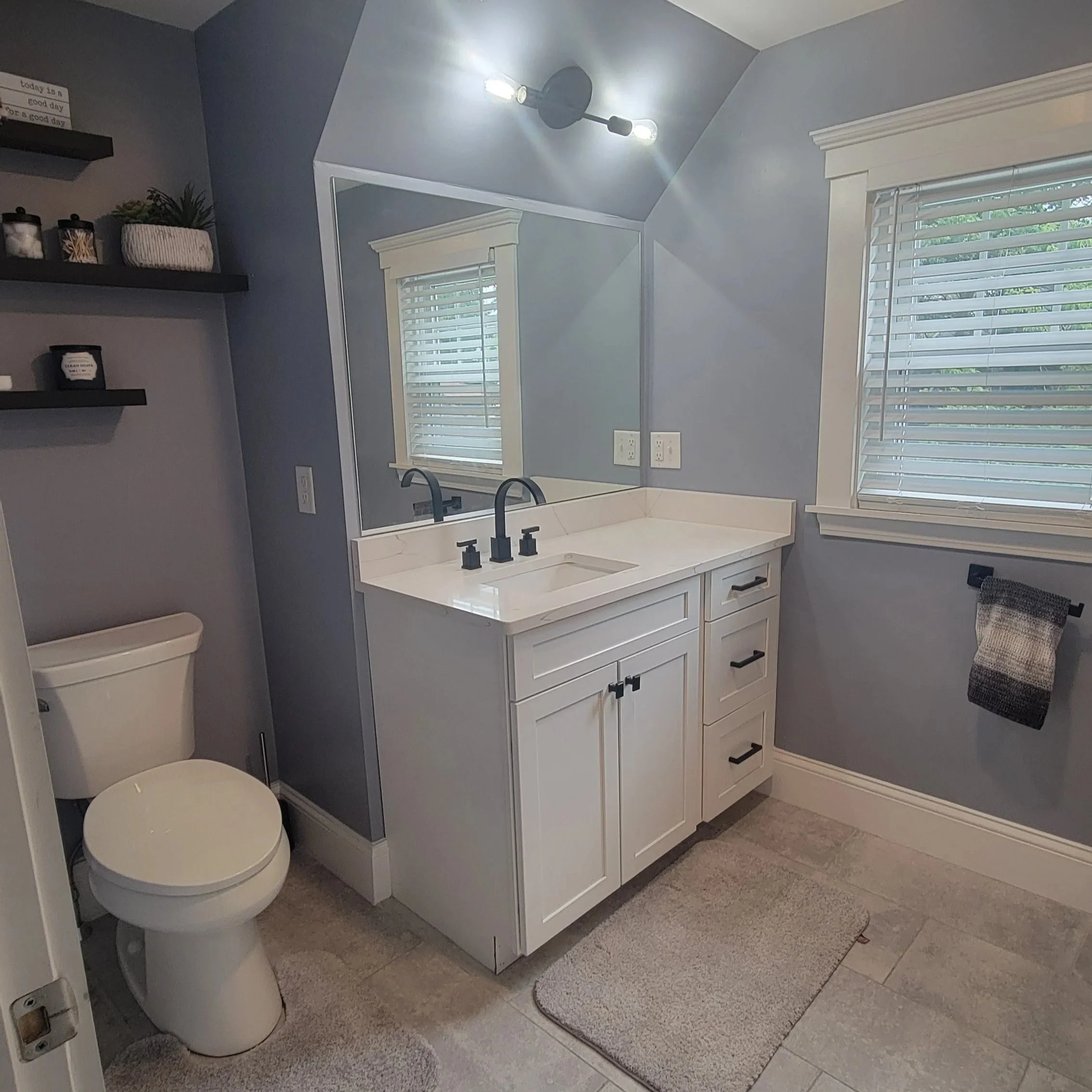 A modern bathroom with a white vanity, black fixtures, and a large mirror. There are two wall-mounted light fixtures above the mirror, and a window with white blinds on the right. A toilet is visible on the left, and decorative items are on black she
