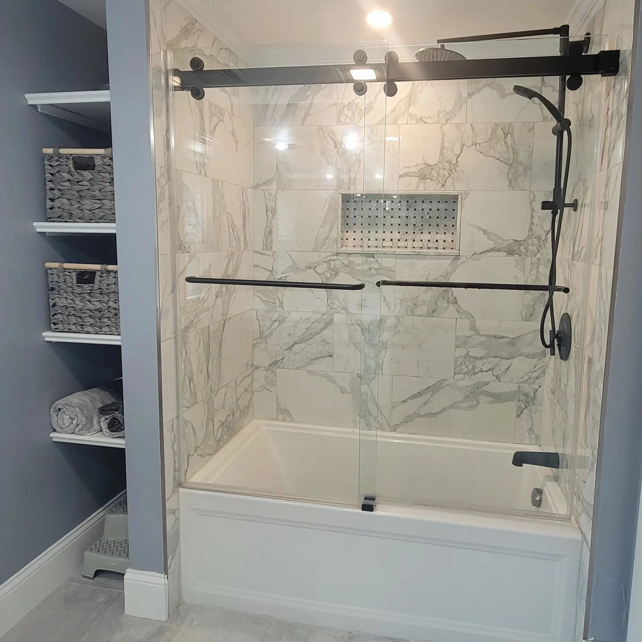 A walk-in shower with marble tiles, a black metal sliding door, a black showerhead, and a built-in niche for toiletries. Renovated by Structure Pro in Massachusetts
