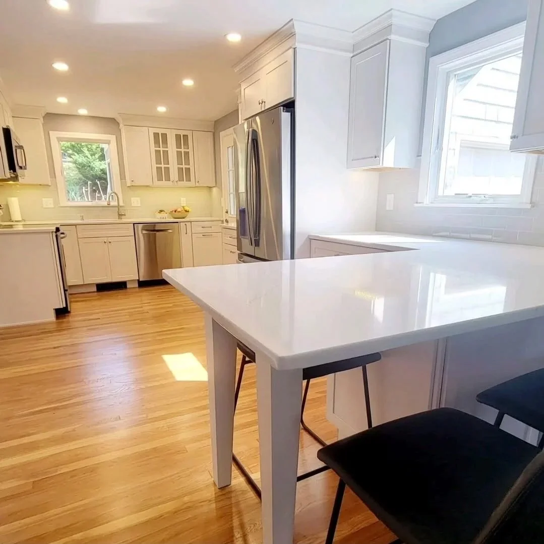 Bright, modern kitchen with white cabinets, stainless steel appliances, hardwood floors, and a white countertop island with black chairs. Renovated Structure Pro in Massachusetts