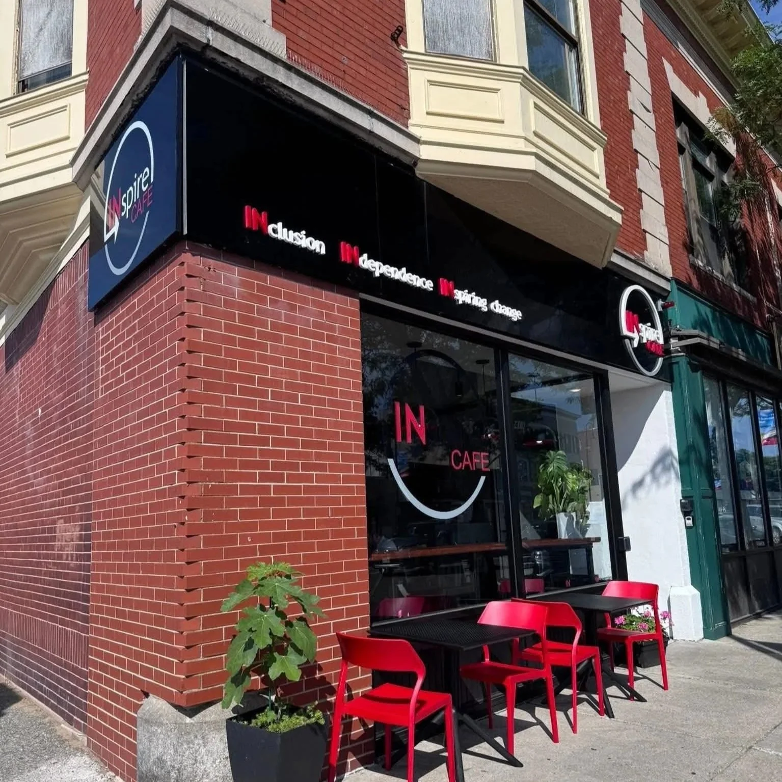 Exterior of modern cafe with red chairs and black tables outside. The cafe has large glass windows and a black sign with white and red lettering that says "Inspiration, Independence, Rising Change." Build by Structure Pro in Massachusetts