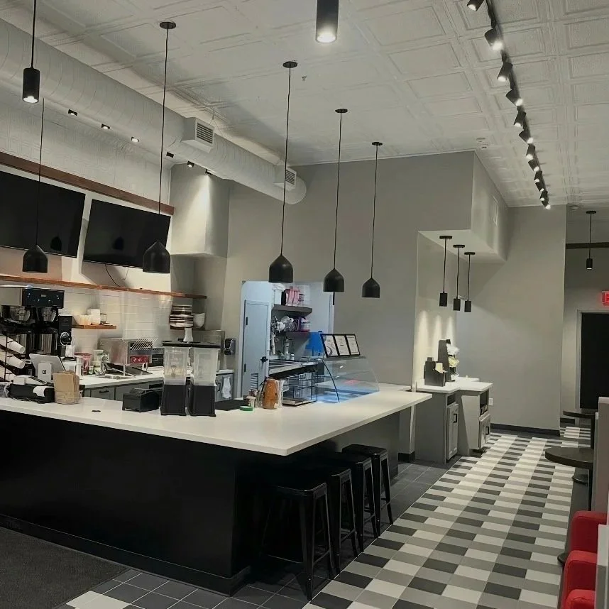 Modern coffee shop interior with a white and black color scheme, four black pendant lights hanging over a counter, coffee-making equipment, and checkered tile floor. Build by Structure Pro in Massachusetts