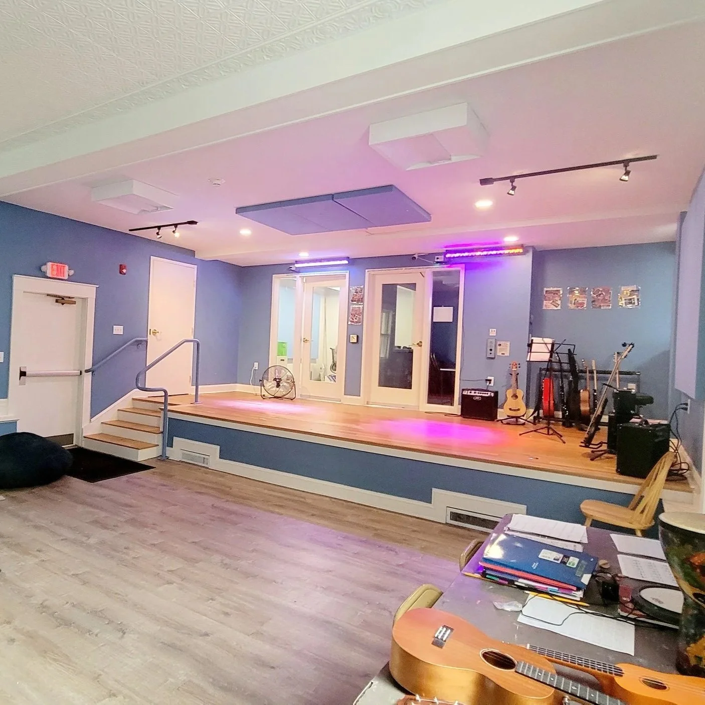 A small stage with musical instruments including guitars, an amp, and a microphone, set up in a room with blue walls and wooden flooring, illuminated with stage lights. Boys and Girls Club building project build by Structure Pro in Massachusetts