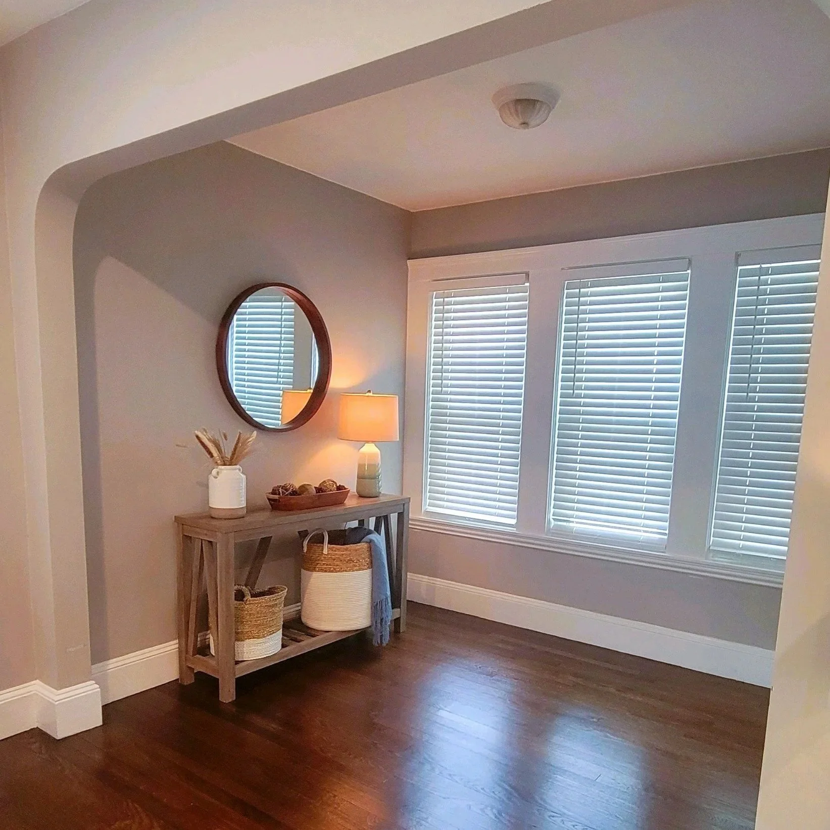 A corner of room with a wooden console table holding a table lamp, decorative bowls, and a white vase with dried plants. A round mirror is above the table. Three windows with white blinds and hardwood floors. Built Structure Pro in Massachusetts