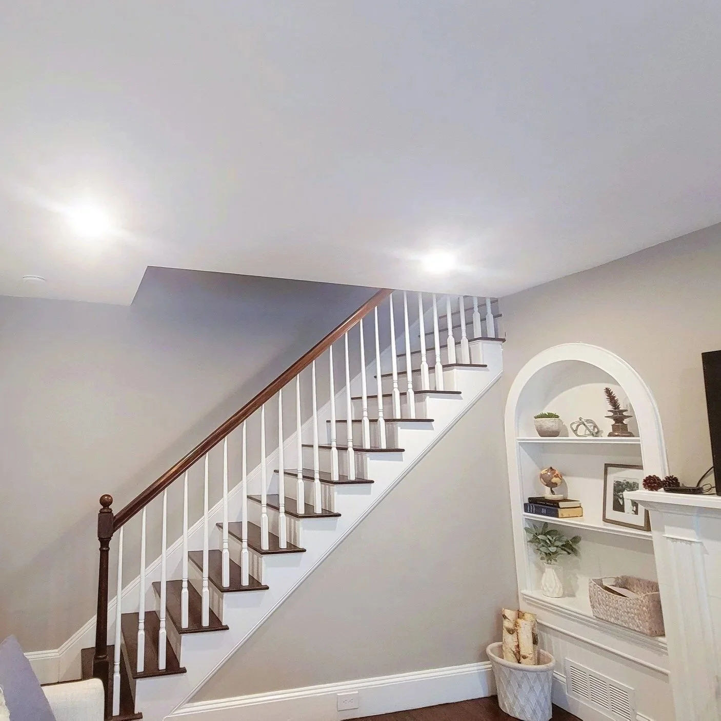 Interior view of a living room corner featuring a staircase with white spindles, dark wood handrail and treads, a gray wall, built-in white shelves with decorative items. Built Structure Pro in Massachusetts.
