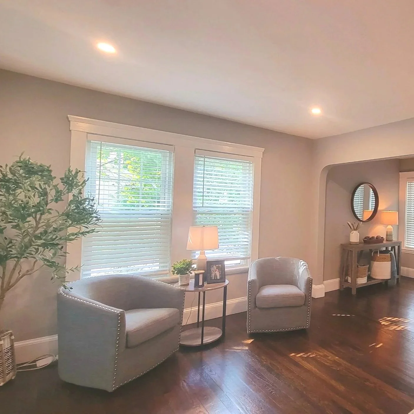 Living room with two beige armchairs, a small side table with a lamp, and a large window with blinds, wooden floor, and a console table in the background with a mirror and decor. Renovated by Structure Pro in Massachusetts