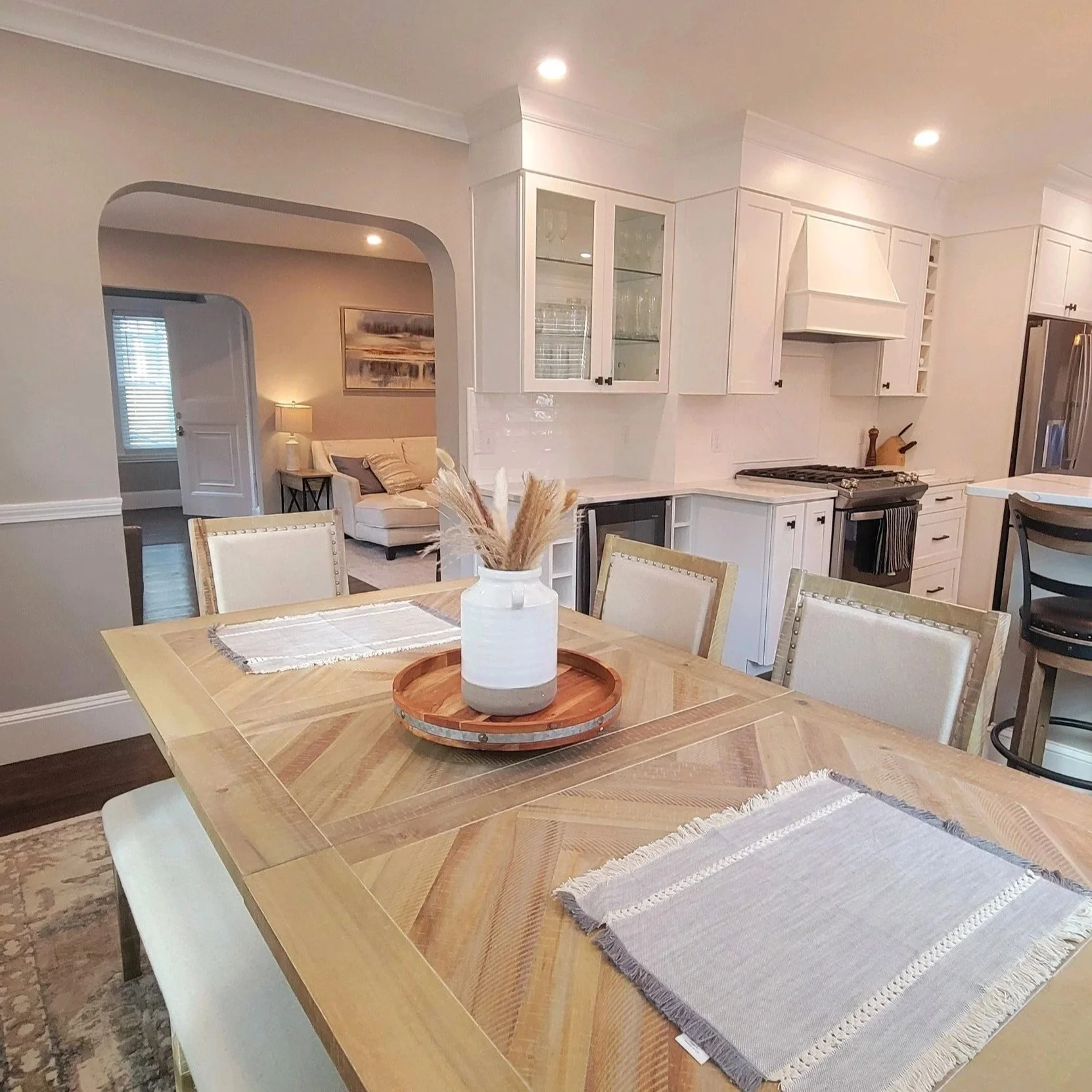 Open-concept kitchen and dining area with a wooden dining table. Features hite cabinets, a stove with a chimney, and a refrigerator, with view into a living room with beige walls, a white sofa, and framed artwork.Built Structure Pro in Massachusetts