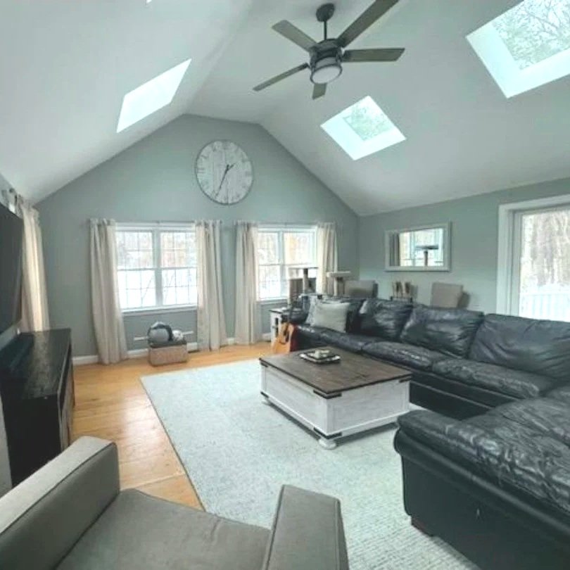 A living room with vaulted ceiling, skylights, and large windows, featuring a black leather sofa, gray armchair, coffee table, and a ceiling fan. Built Structure Pro in Massachusetts