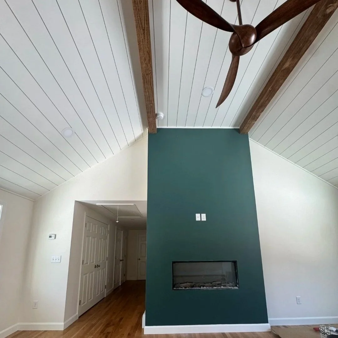 Living room interior with a teal accent wall and built-in fireplace, white paneled vaulted ceiling, wooden beams, and a ceiling fan, with hardwood flooring and a hallway with double doors. Built Structure Pro in Massachusetts