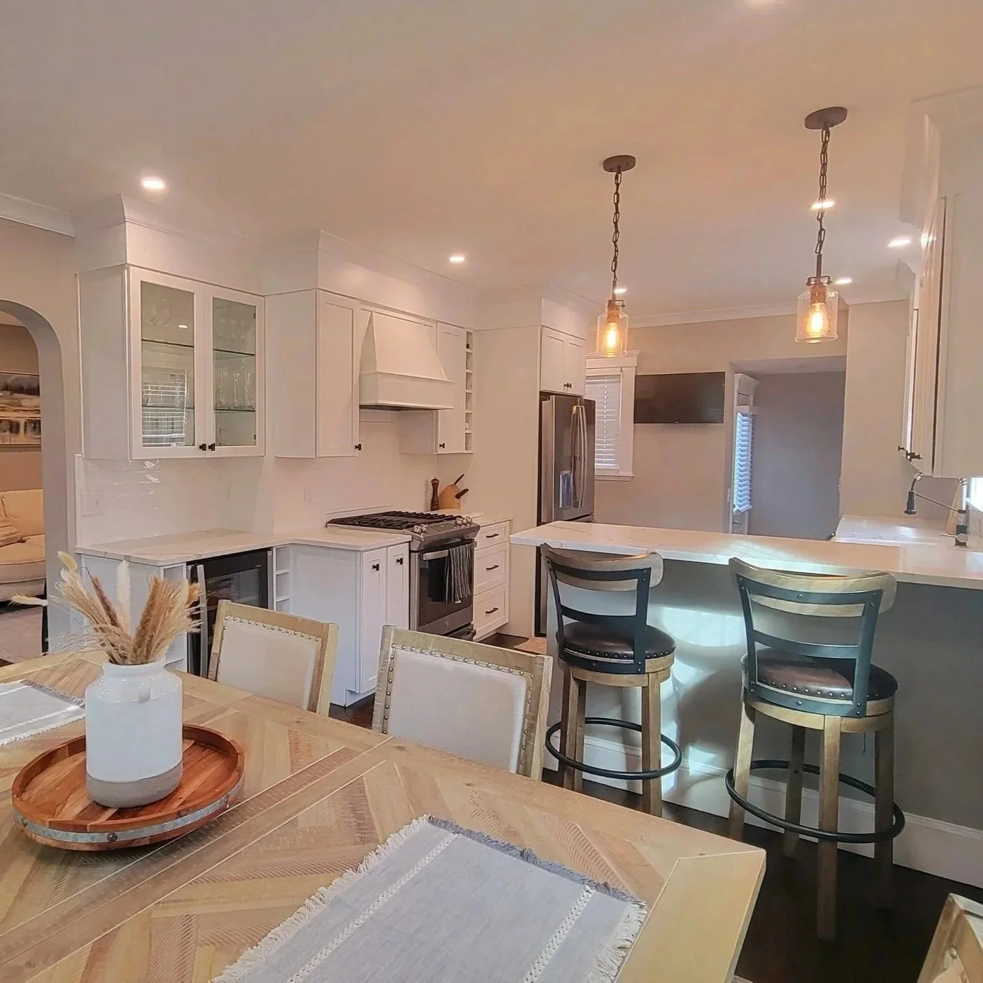 Modern white kitchen with island, pendant lighting, and bar stools, adjacent to dining area with a vase and placemats on a wooden table. Renovated Structure Pro in Massachusetts