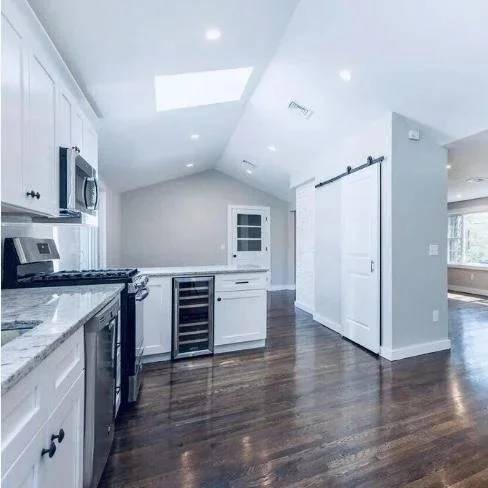 Modern kitchen with white cabinets, stainless steel appliances, a wine cooler, and a white sliding barn door, with hardwood floors and a sloped ceiling with skylights. Renovated Structure Pro in Massachusetts