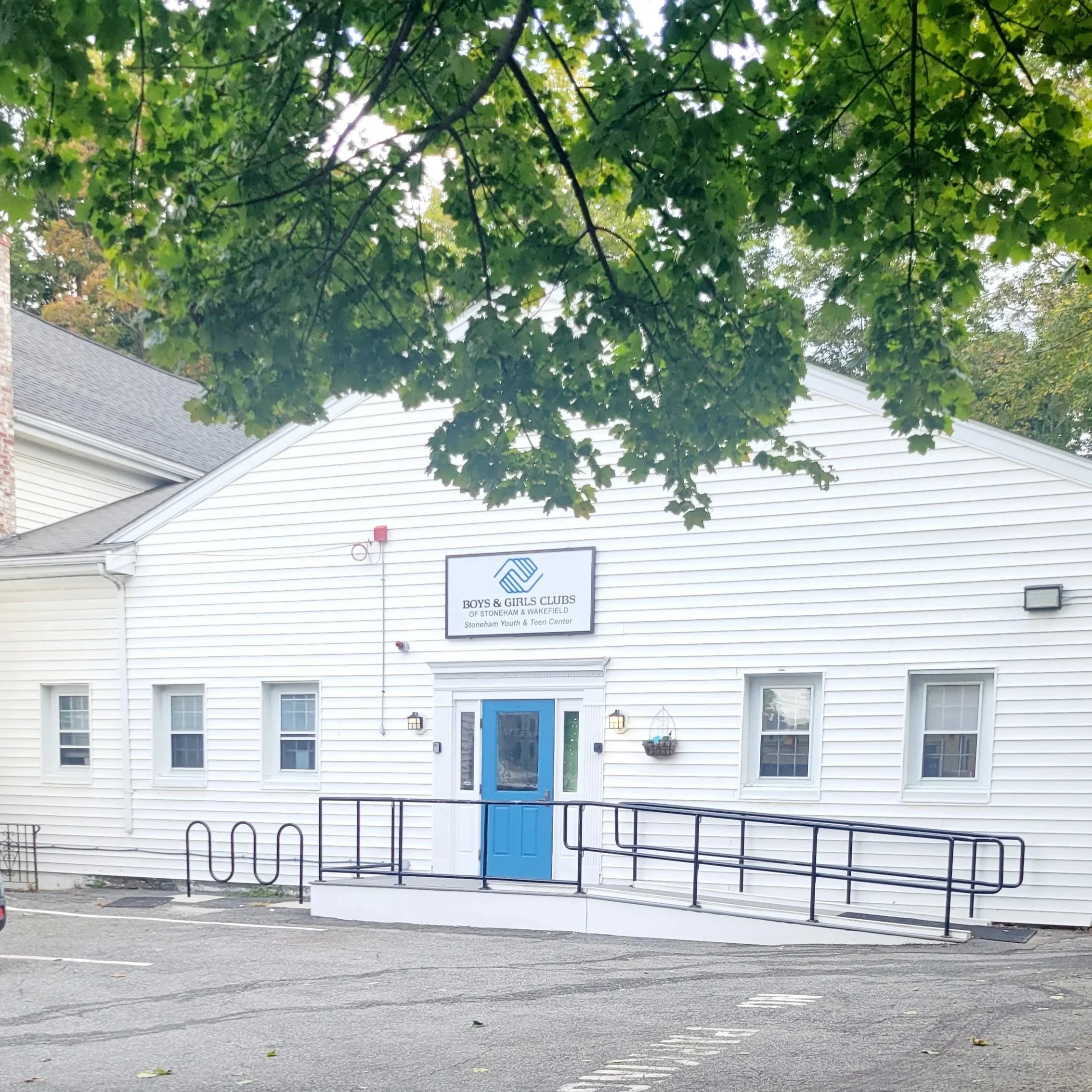 Front view of a white building with a blue door, sign above door reads 'Boys & Girls Clubs,' with trees and parked cars in the foreground. Wheelchair ADA accesible ramp built by Structure Pro in Massachusetts