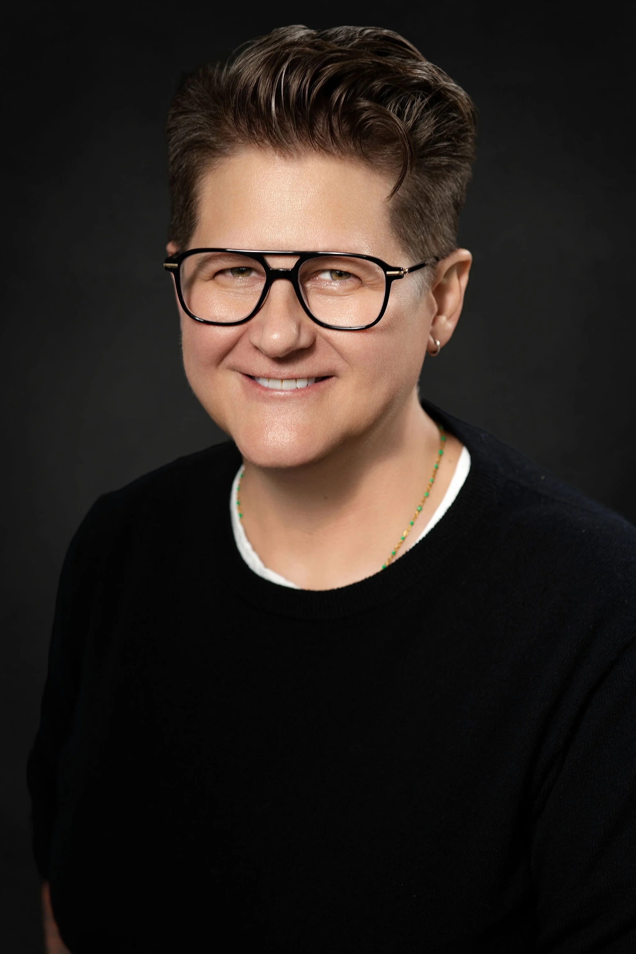 A smiling person with short brown hair, wearing black glasses, a black sweater, and a beaded necklace, posed against a dark background.