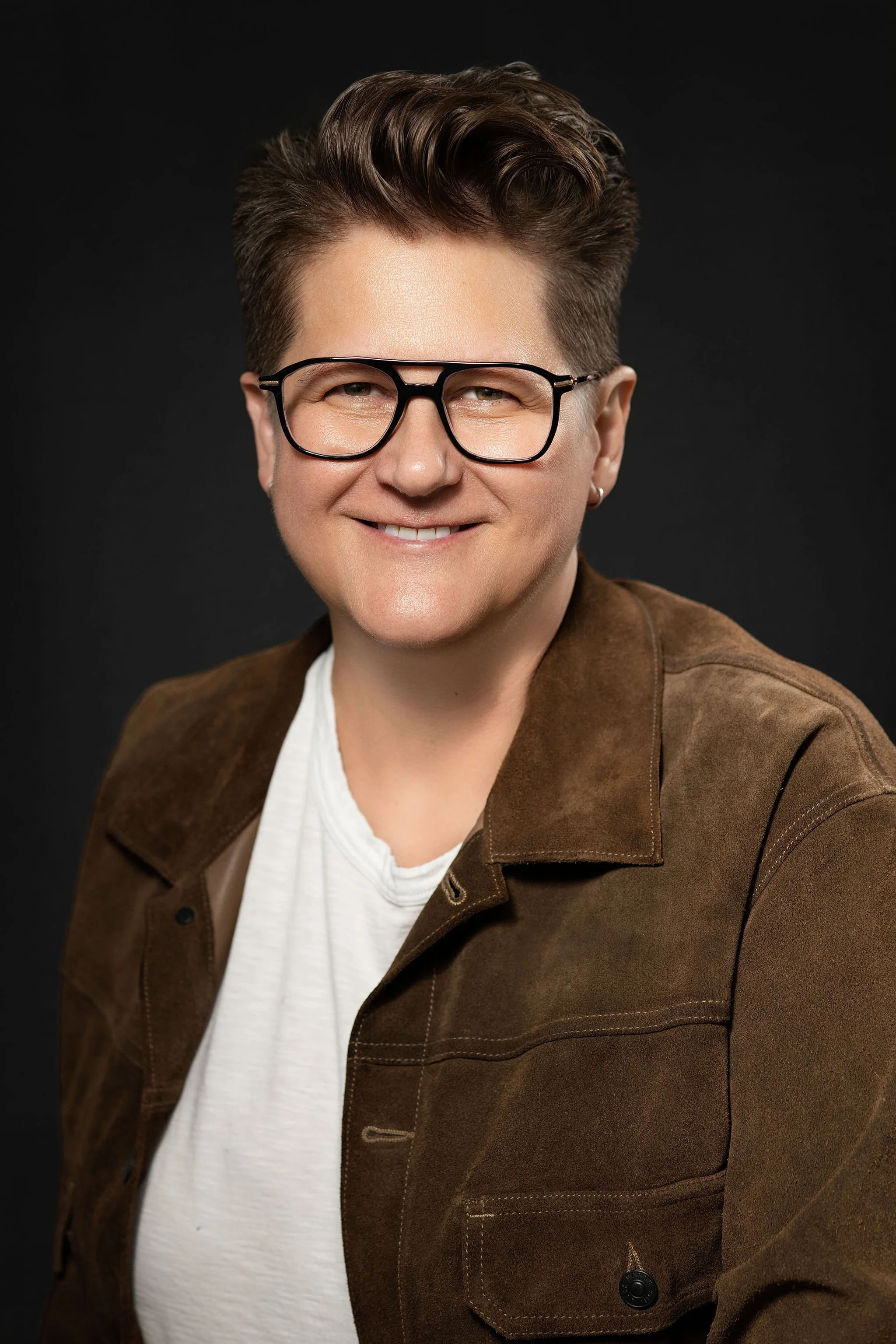 Portrait of a smiling woman with short brown hair, wearing large glasses, a white t-shirt, and a brown jacket, against a dark background.