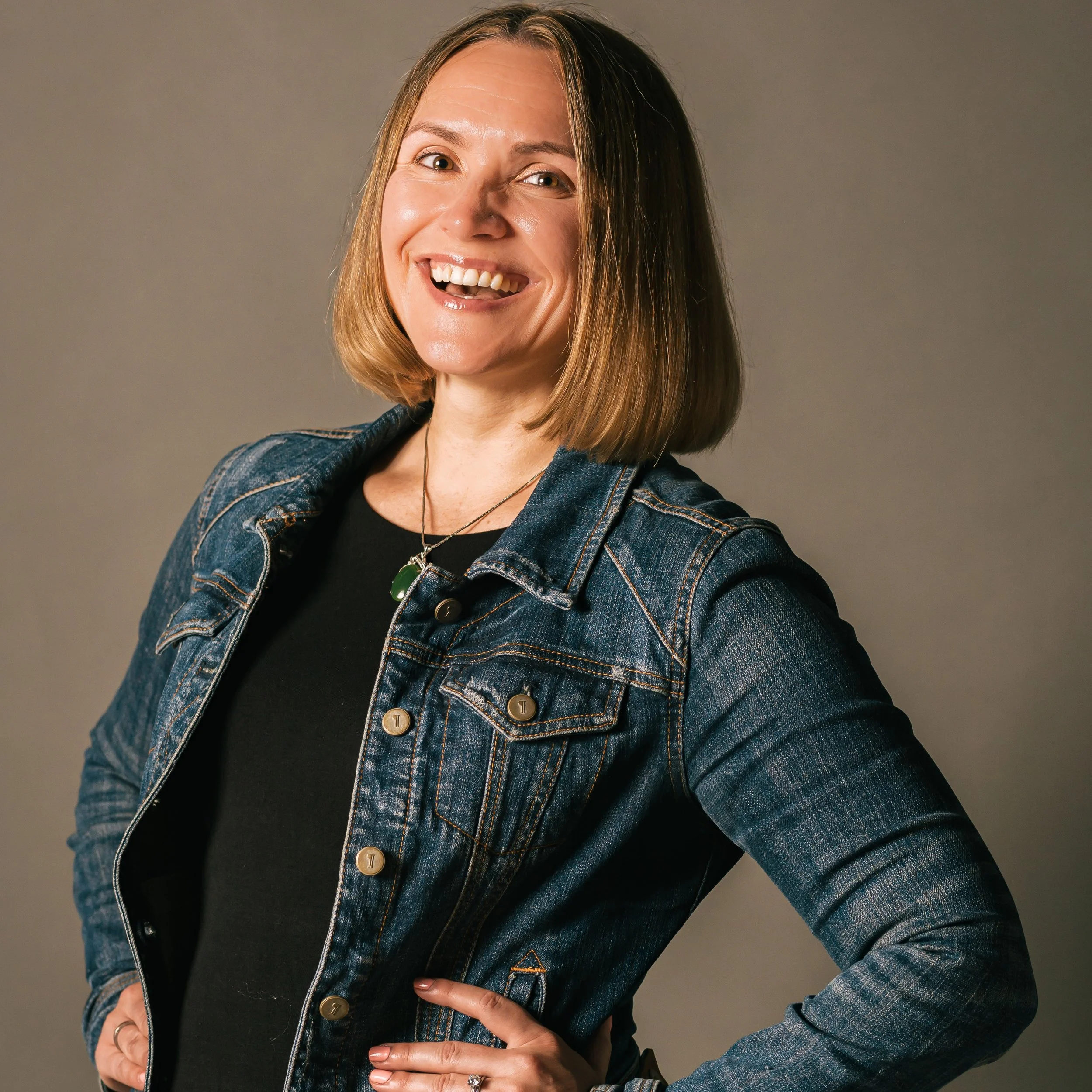 A woman, our founder, Kathleen,  with shoulder-length blonde hair wearing a denim jacket and black shirt, standing against a plain background.
