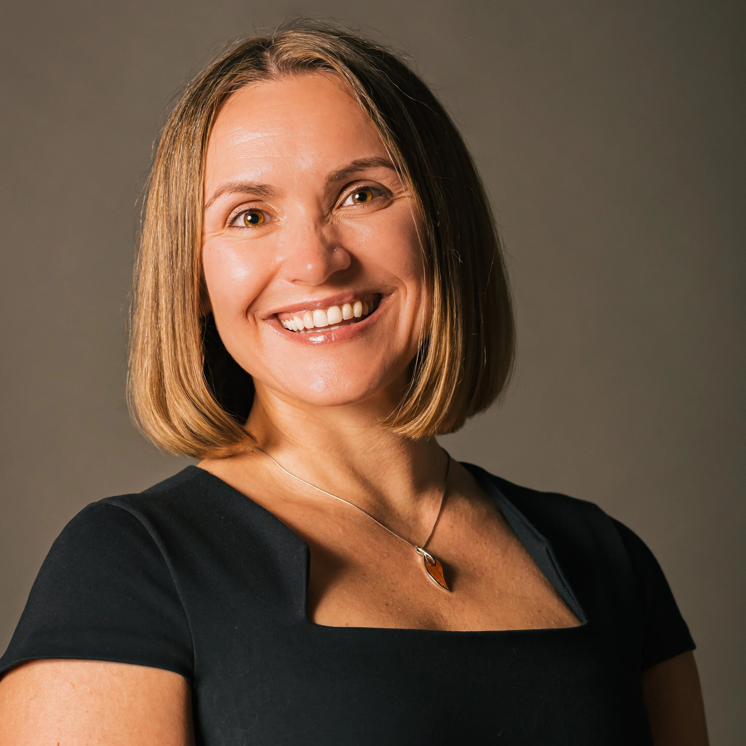 Portrait of a woman, our founder, Kathleen, with shoulder-length brown hair, wearing a black top and a pendant necklace, against a plain gray background.