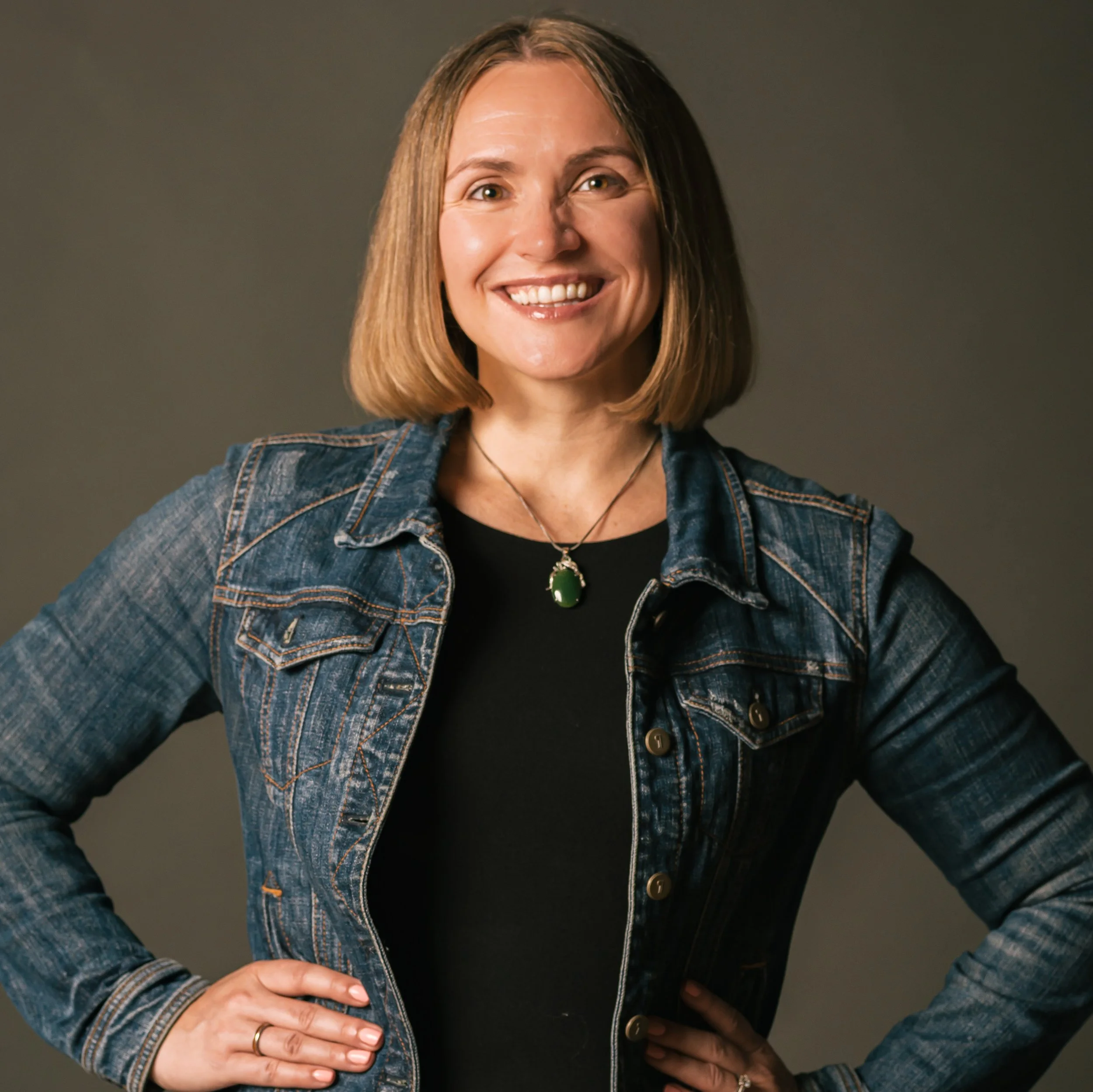 A woman, our founder, Kathleen, with shoulder-length light brown hair smiling, wearing a denim jacket over a black top, and a green pendant necklace, standing with hands on hips against a gray background.