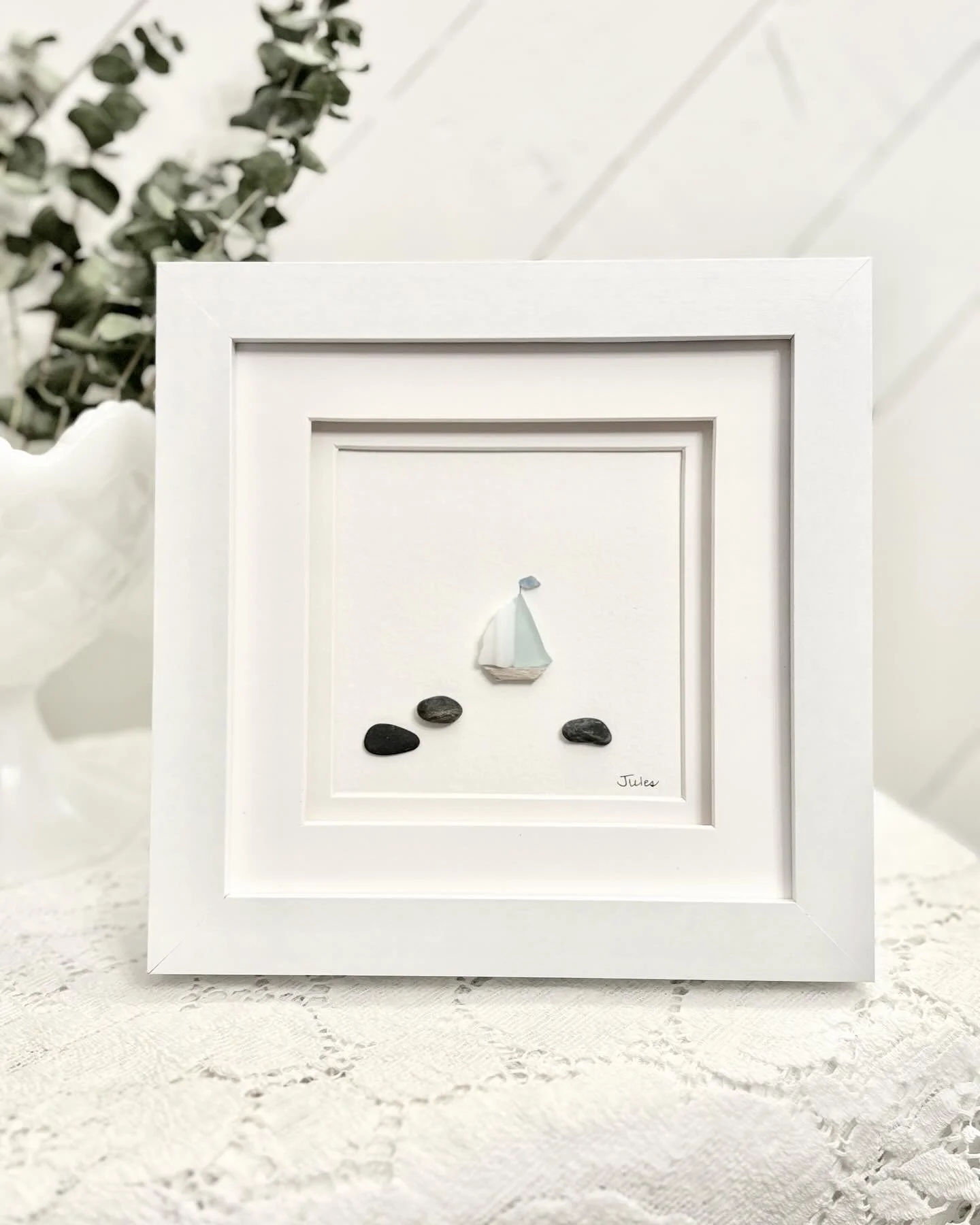 Framed artwork of a sailboat made with sea glass and stones, positioned on a white lace tablecloth with green foliage in the background.