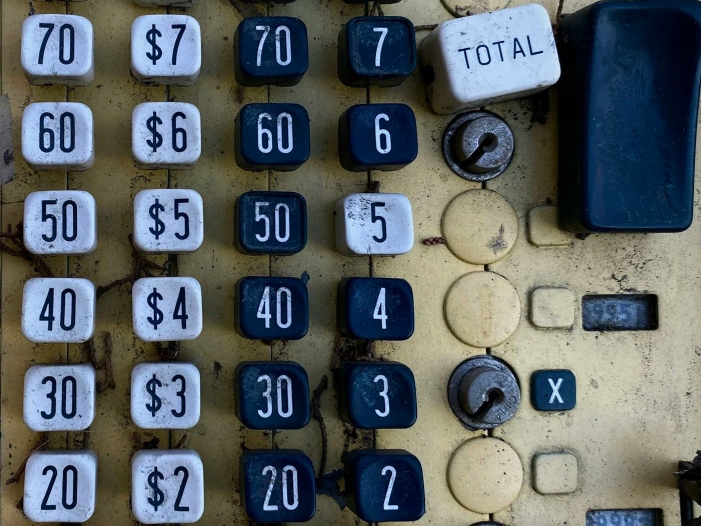 Close-up of an old, weathered mechanical calculator or cash register with buttons for various dollar amounts and operations, including a total and an X button, with a lock switch and compartments.