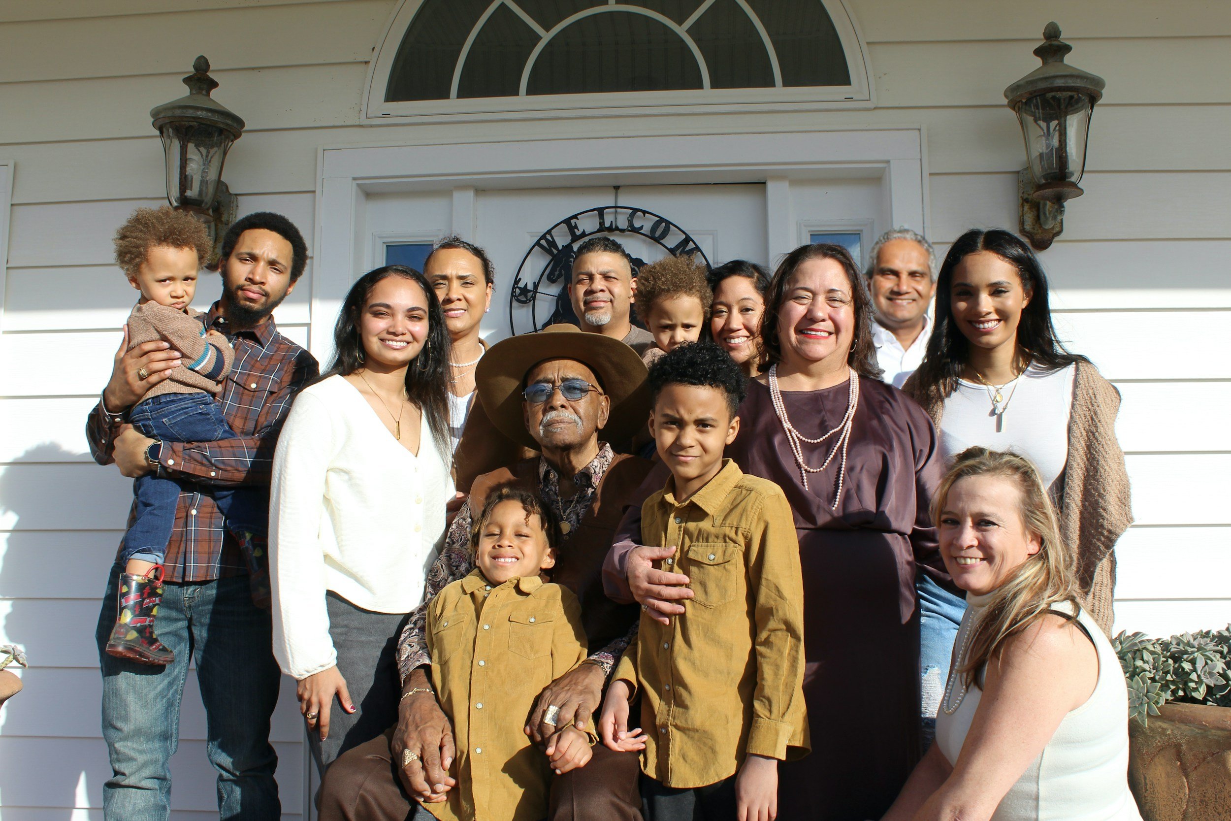 A large multigenerational family family standing outside a house, posing for a group photo. The group includes children, adults, and seniors, all smiling and facing the camera.