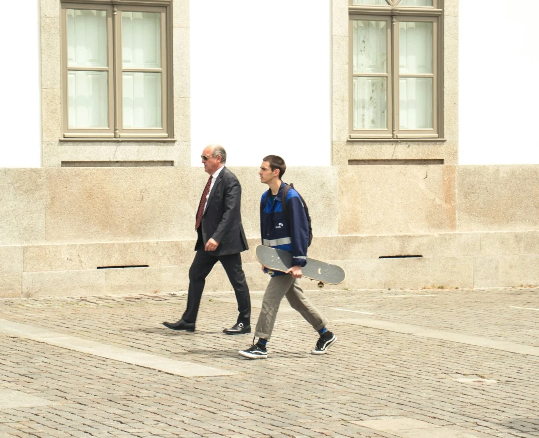 A young man carrying a skateboard walking past an older man in a suit on a cobblestone sidewalk in front of a building with large windows.