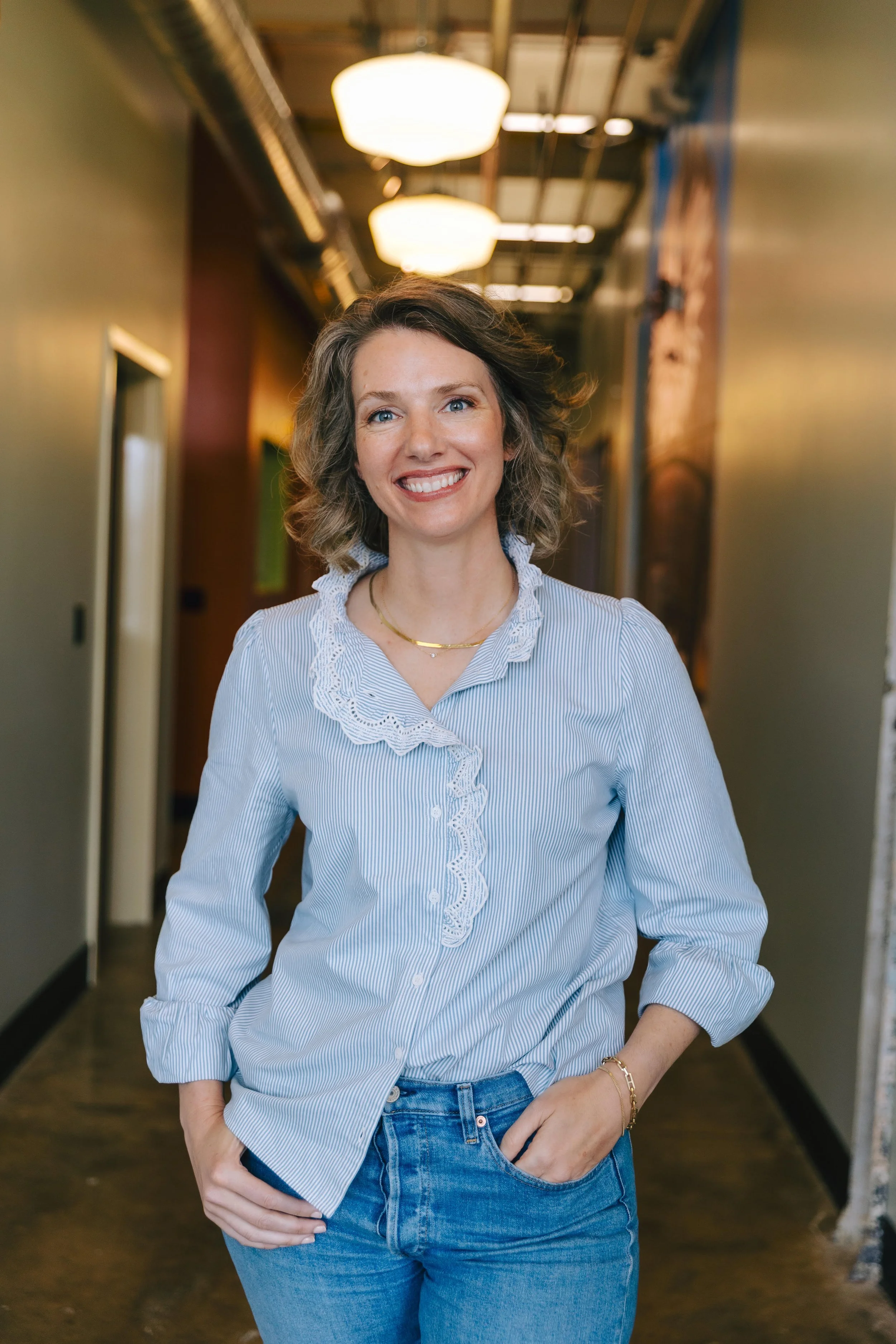 A photo of Laurel-- a friendly-looking, professional Caucasian woman with brown hair-- smiling, wearing a light blue striped blouse with ruffles and jeans, standing in a hallway with warm lighting.