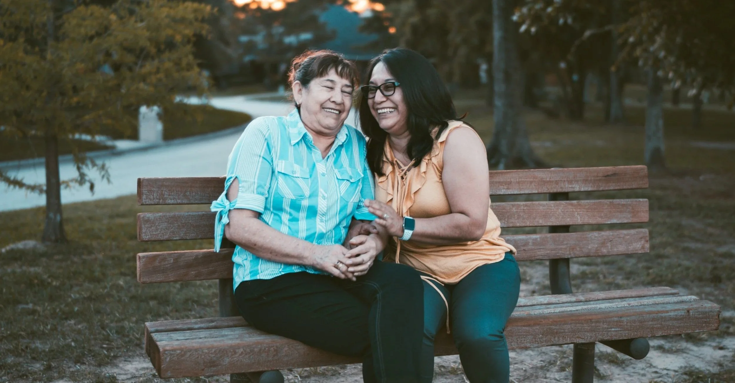 Two women sitting on a wooden park bench, laughing and sharing a joyful moment, surrounded by trees and a walking path.