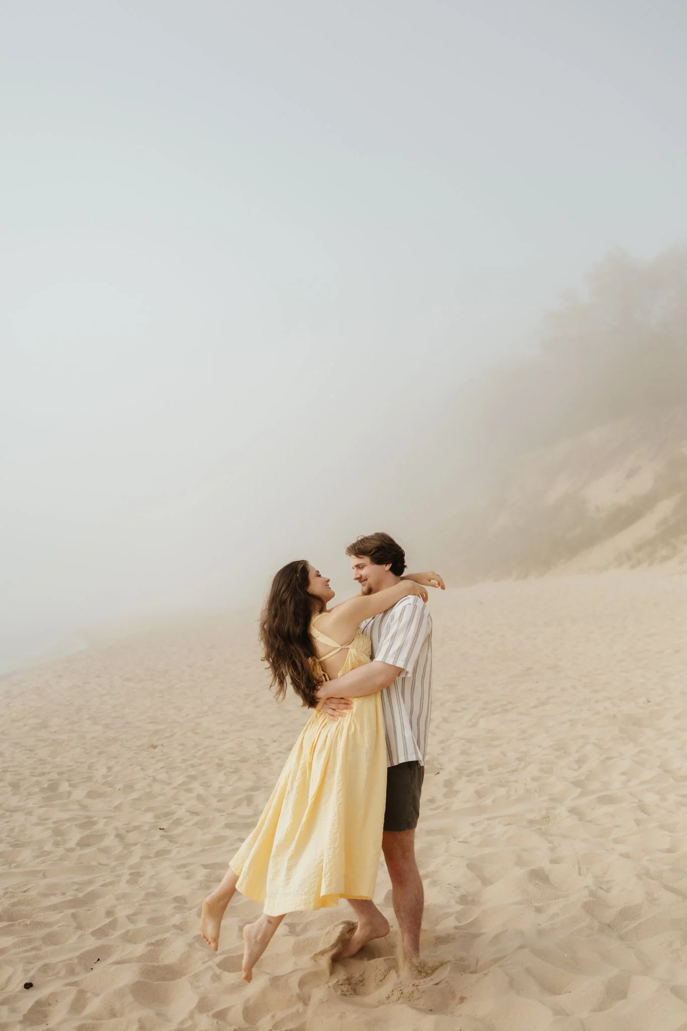 Couple portraits at beach location along Lake Michigan with fog and fun running moment, engagement and proposal.
