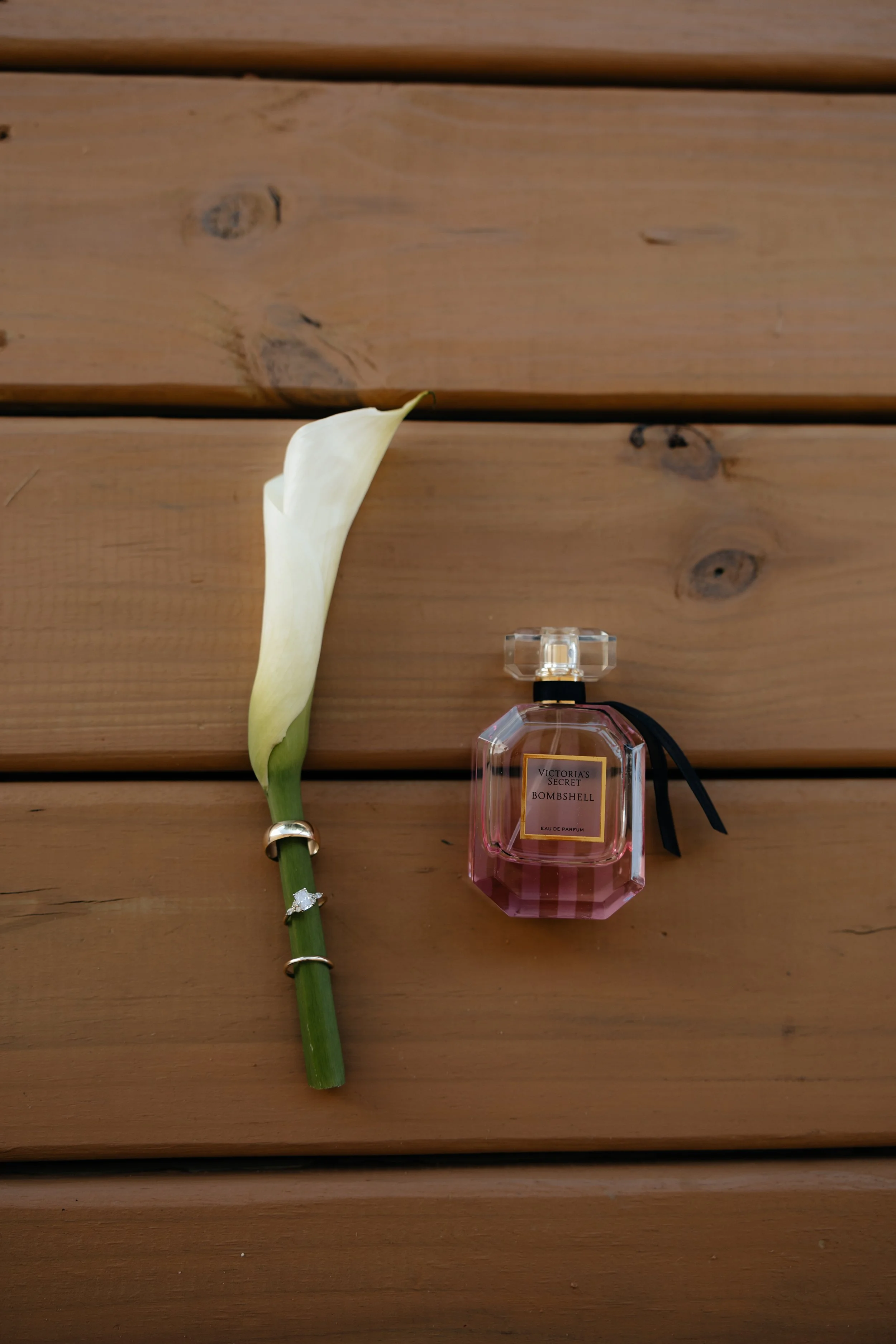 A white calla lily flower with three rings around its stem, positioned next to a pink Victoria's Secret Bombshell perfume bottle on a wooden surface, wedding details simple and elegant.