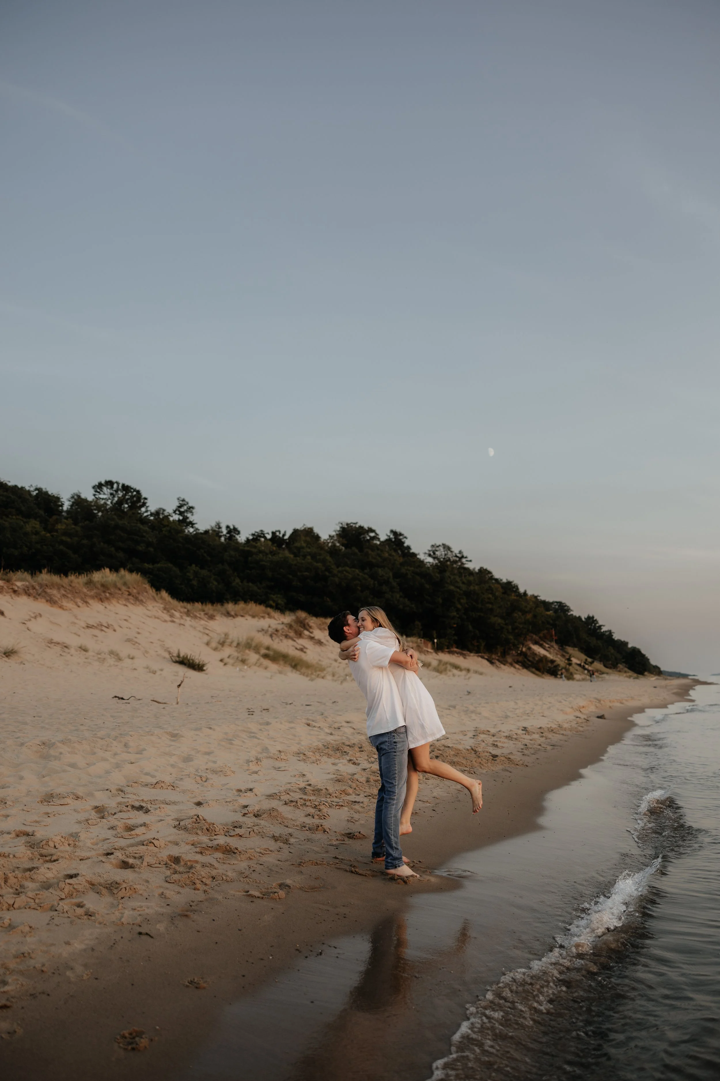 An engaged couple hugging wearing rings at Rosy Mound beach at sunset, with a wooded hillside in the background and the moon visible in the sky.