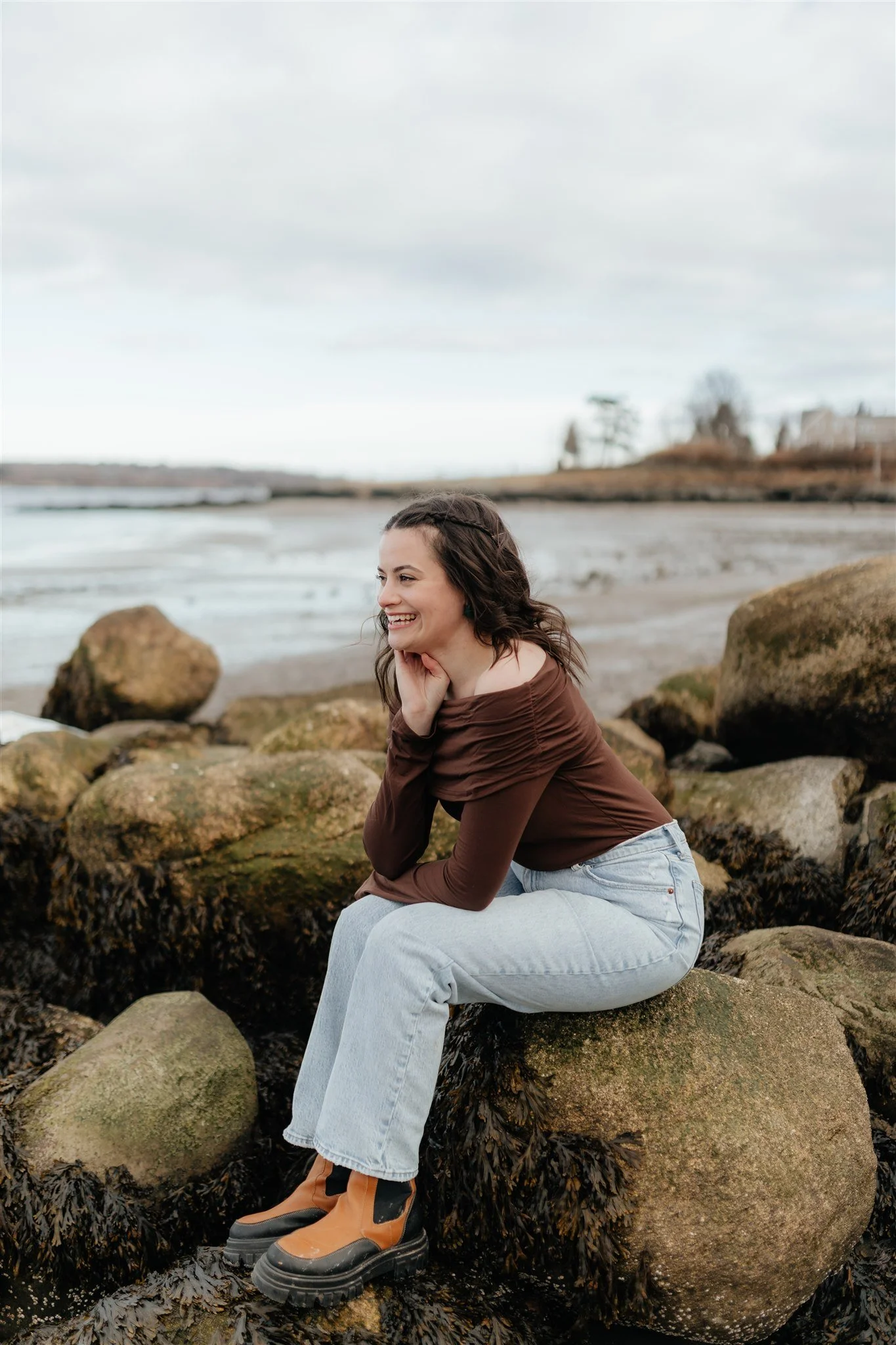 A woman sitting on rocks by the water on the coast of Maine for branding portraits, smiling and looking to the side, wearing a brown top, light jeans, and orange and black boots with a cloudy sky in the background.