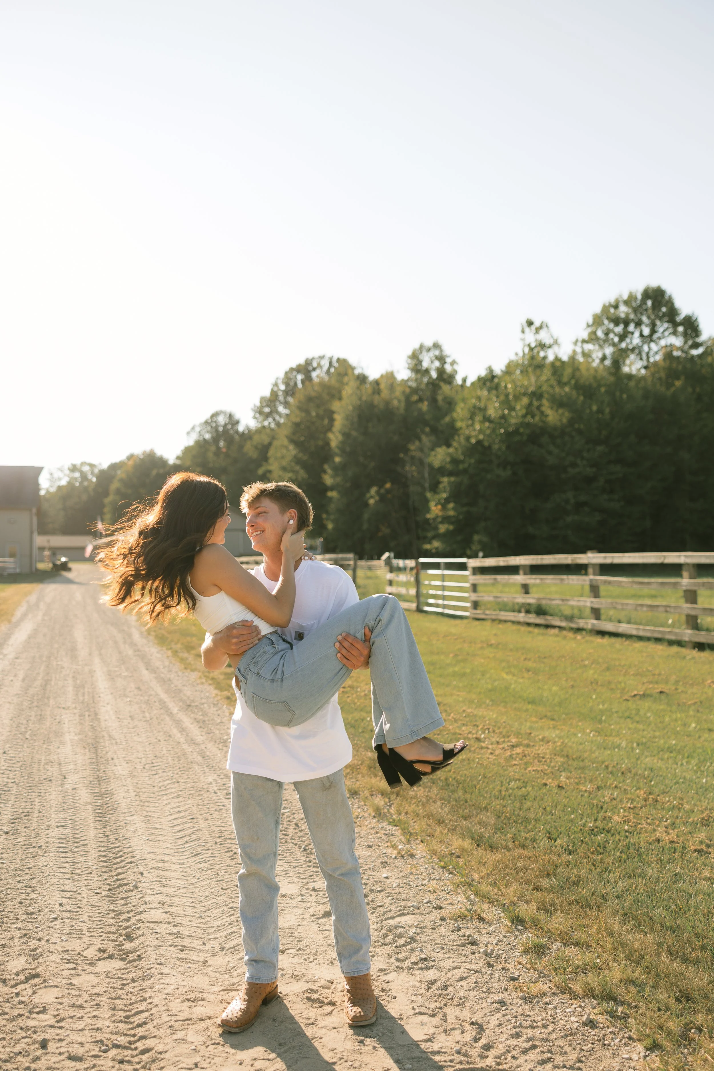 Fun poses for engaged couples on a dirt path in a rural setting with green trees and a wooden fence in the background. They are smiling at each other during sunset.