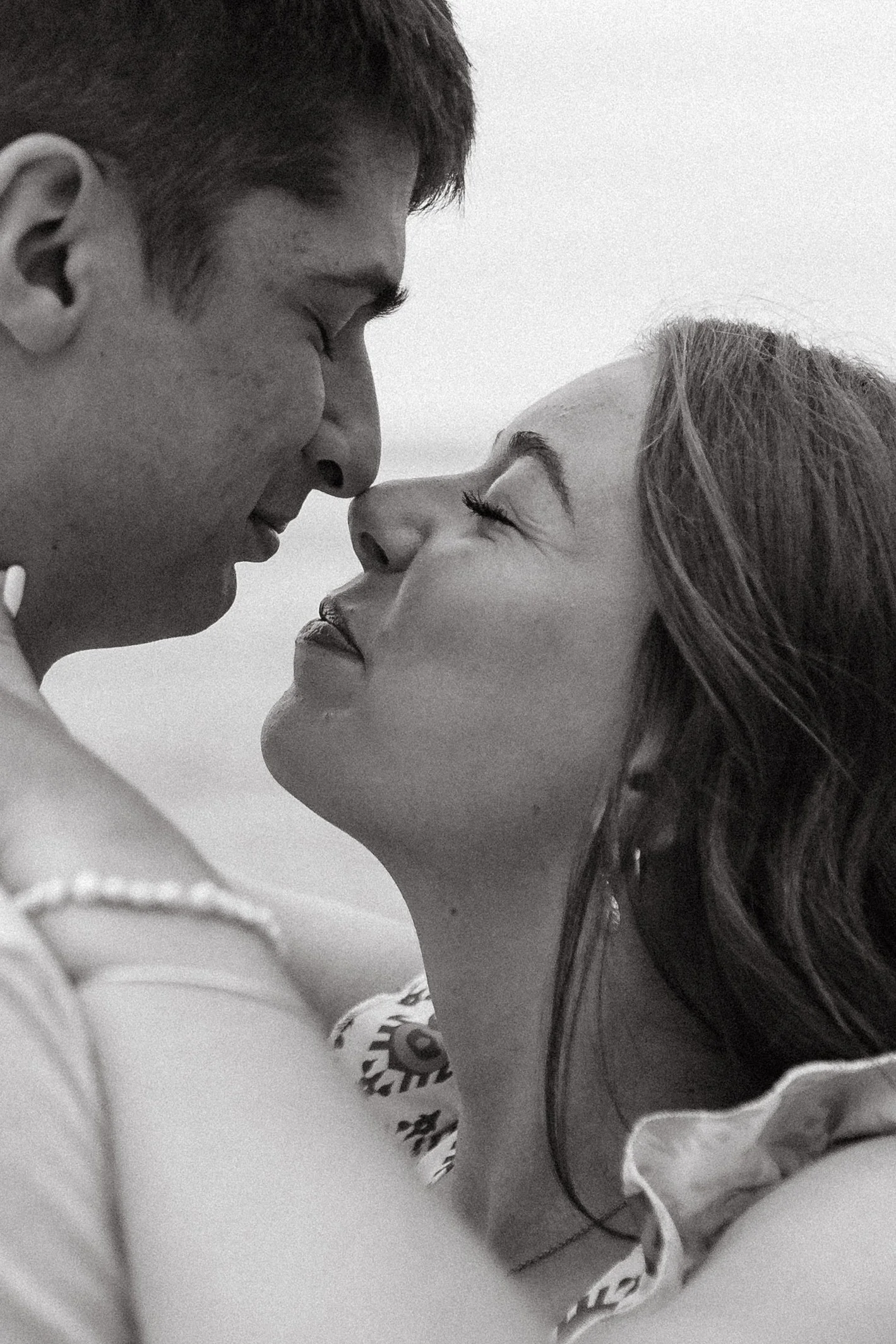 A black and white photograph of a couple in an intimate moment, face to face, with their foreheads and noses touching, smiling softly on the beach of Lake Michigan golden hour.