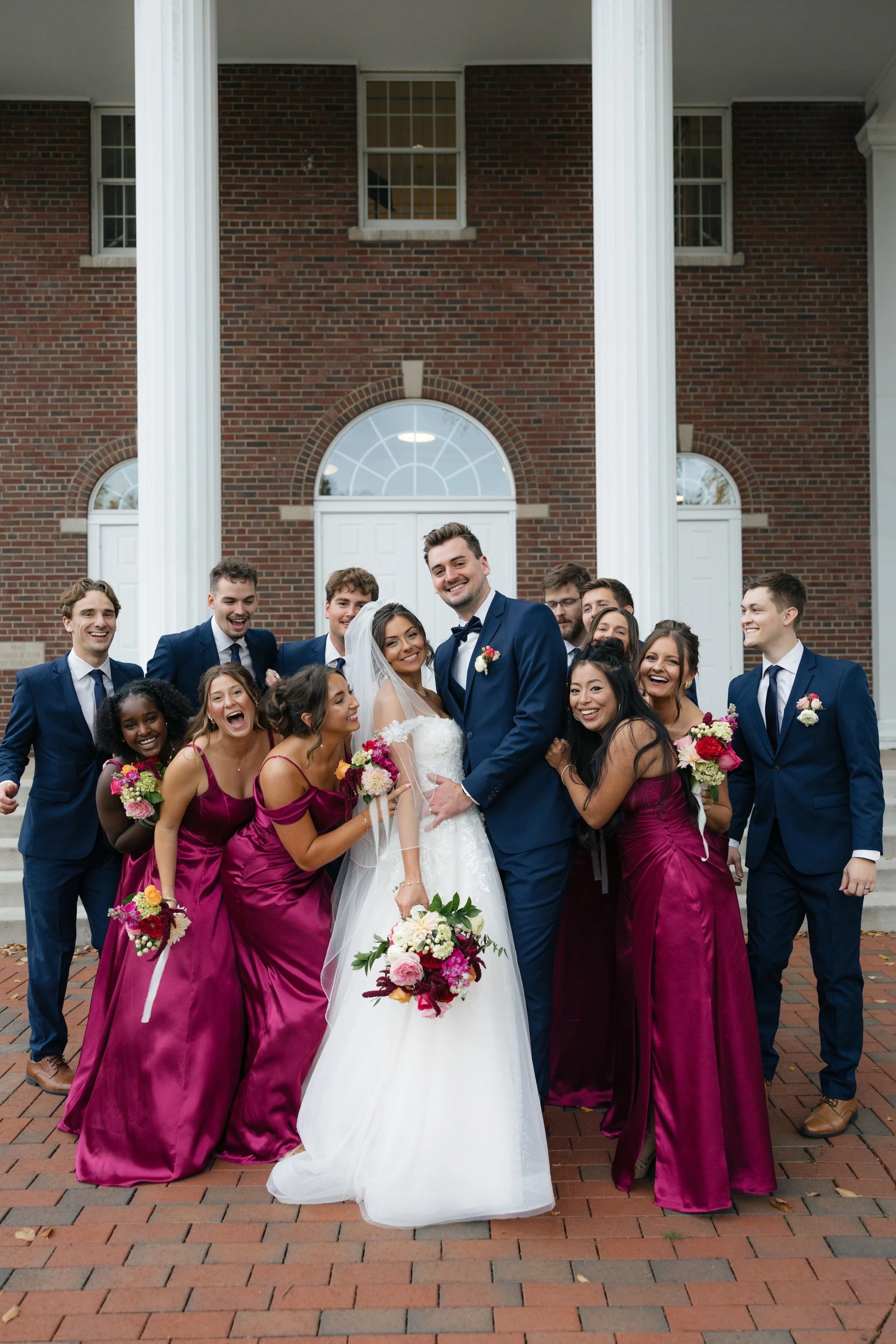 A group of people, including a bride and groom, pose for a photo in front of a brick building with white columns. The bride is in a wedding dress holding a bouquet, and the groom is in a dark blue suit. The group is dressed in formal attire, with wom