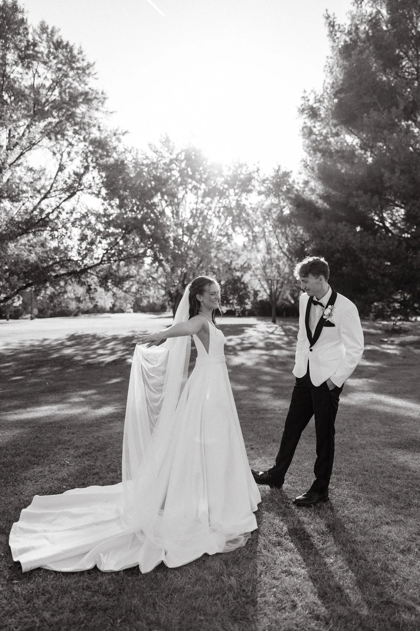 A bride in a wedding gown and veil with groom in a tuxedo with a boutonniere, outdoors on a sunny day with trees in the background at wedding venue in West Michigan.