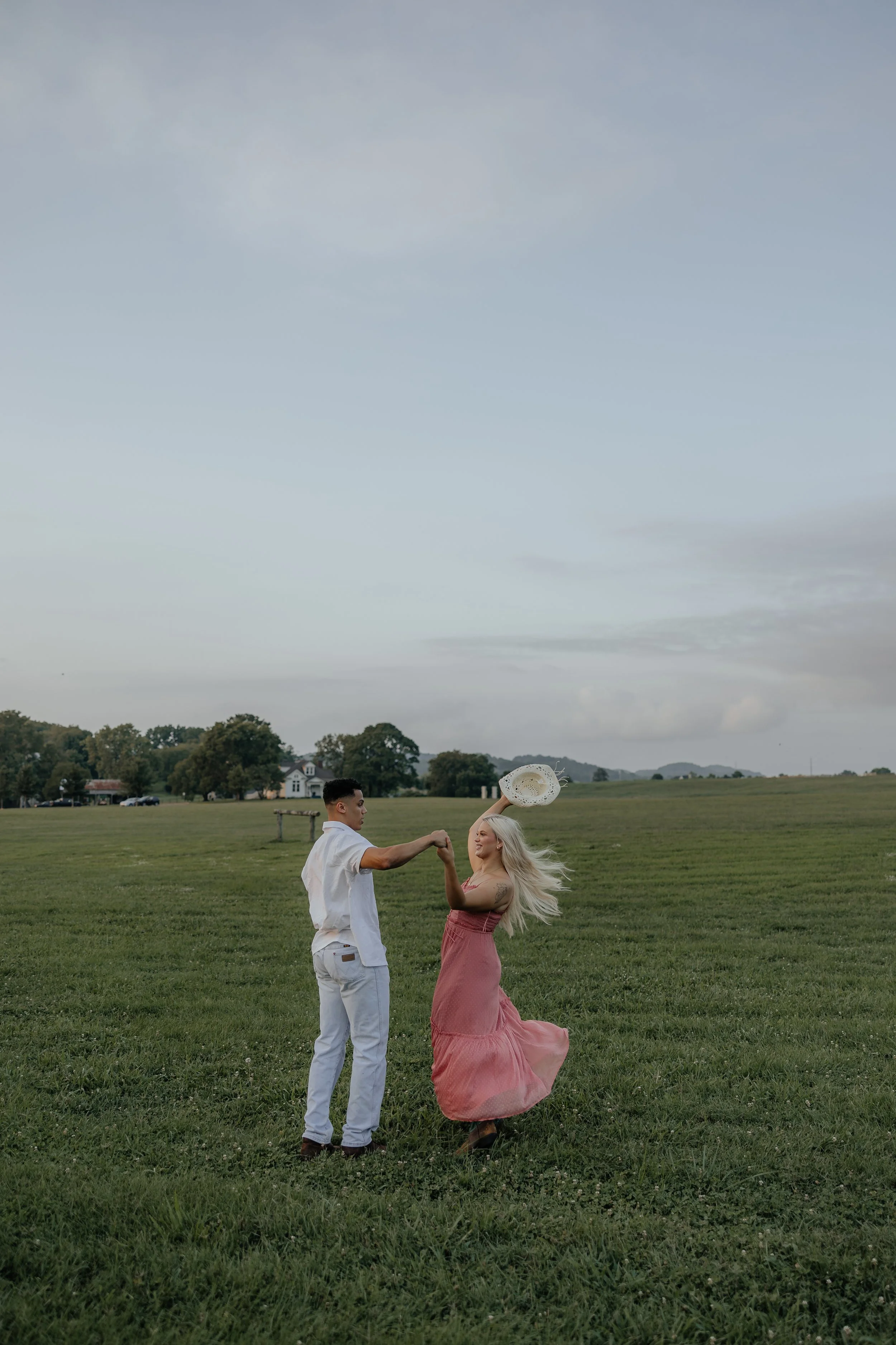 A man and woman dancing in an open grassy field, in Nashville, Tennessee wearing country boots in front of mountain.