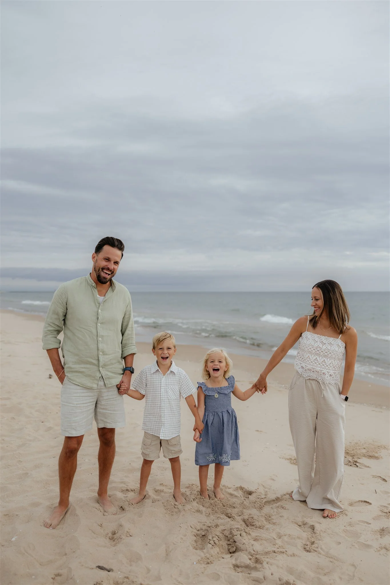 A family of four with two children, holding hands and smiling on a sandy beach with Lake Michigan and cloudy sky in the background.