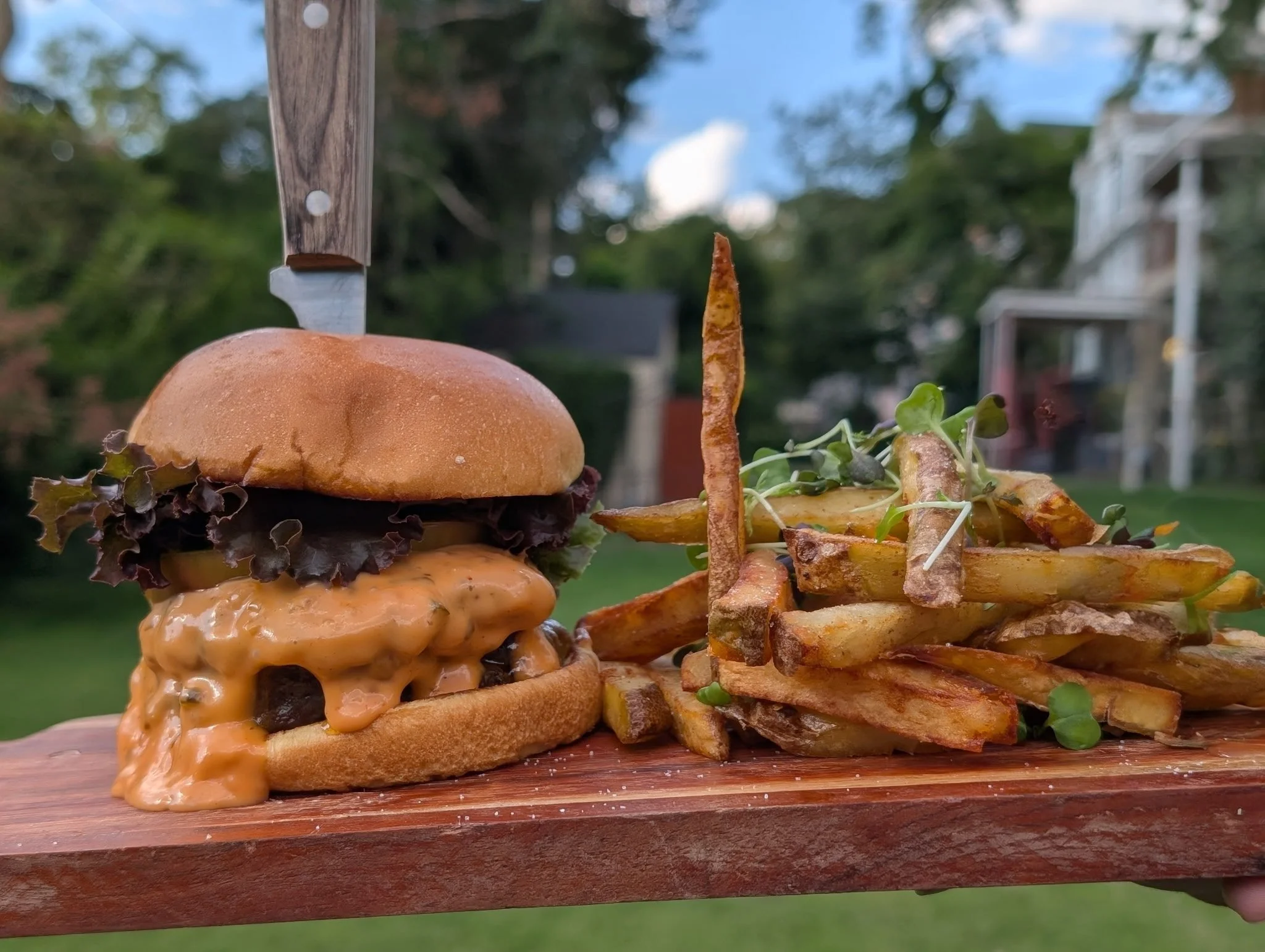 Cheeseburger with lettuce and a knife stabbed through the top bun, served with fries and microgreens on a wooden board outdoors.