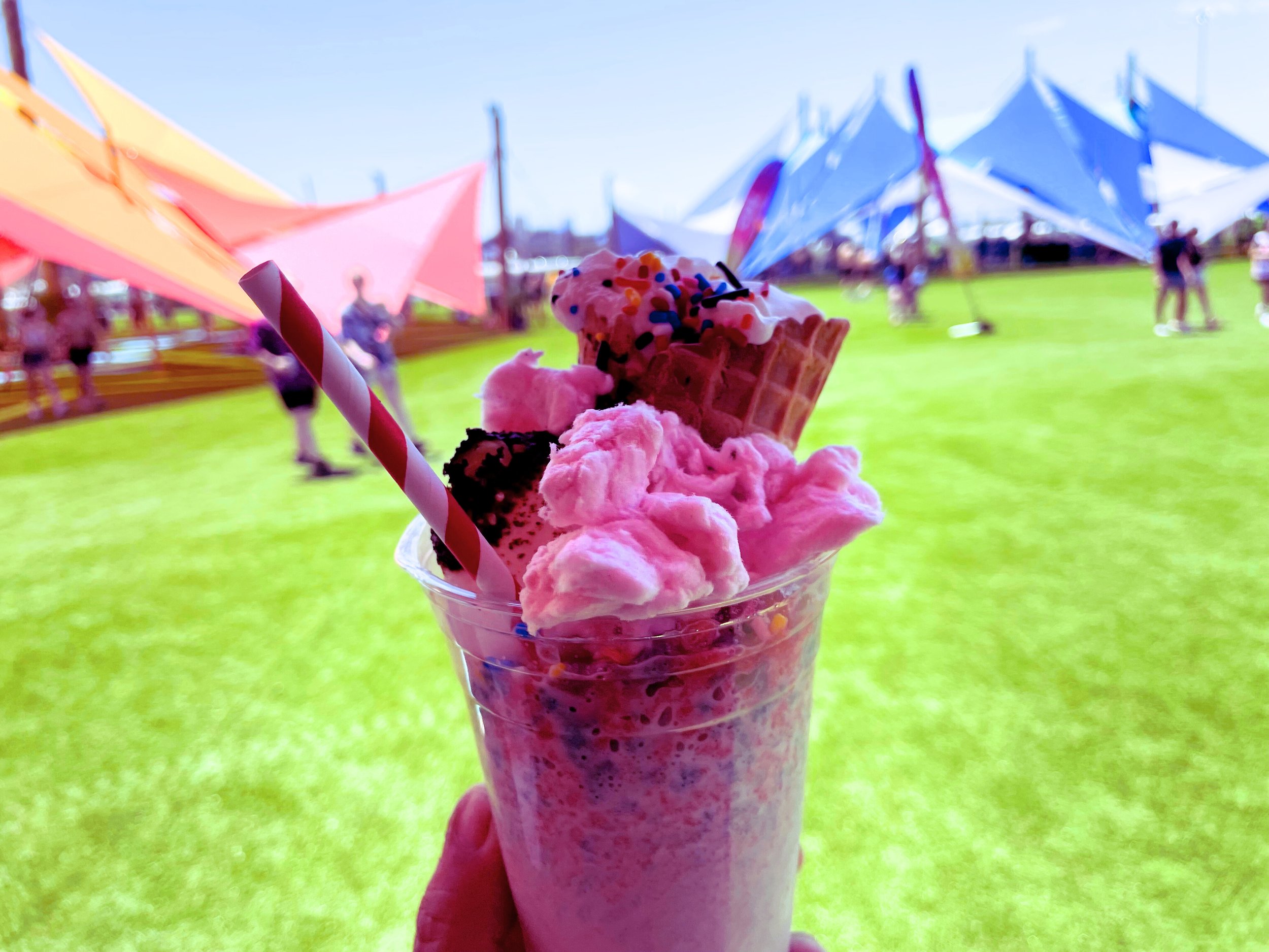 A colorful ice cream float with pink ice cream, rainbow sprinkles, chocolate syrup, a waffle cone, and a striped straw, held up against a vibrant outdoor festival scene with large colorful tents and people in the background.