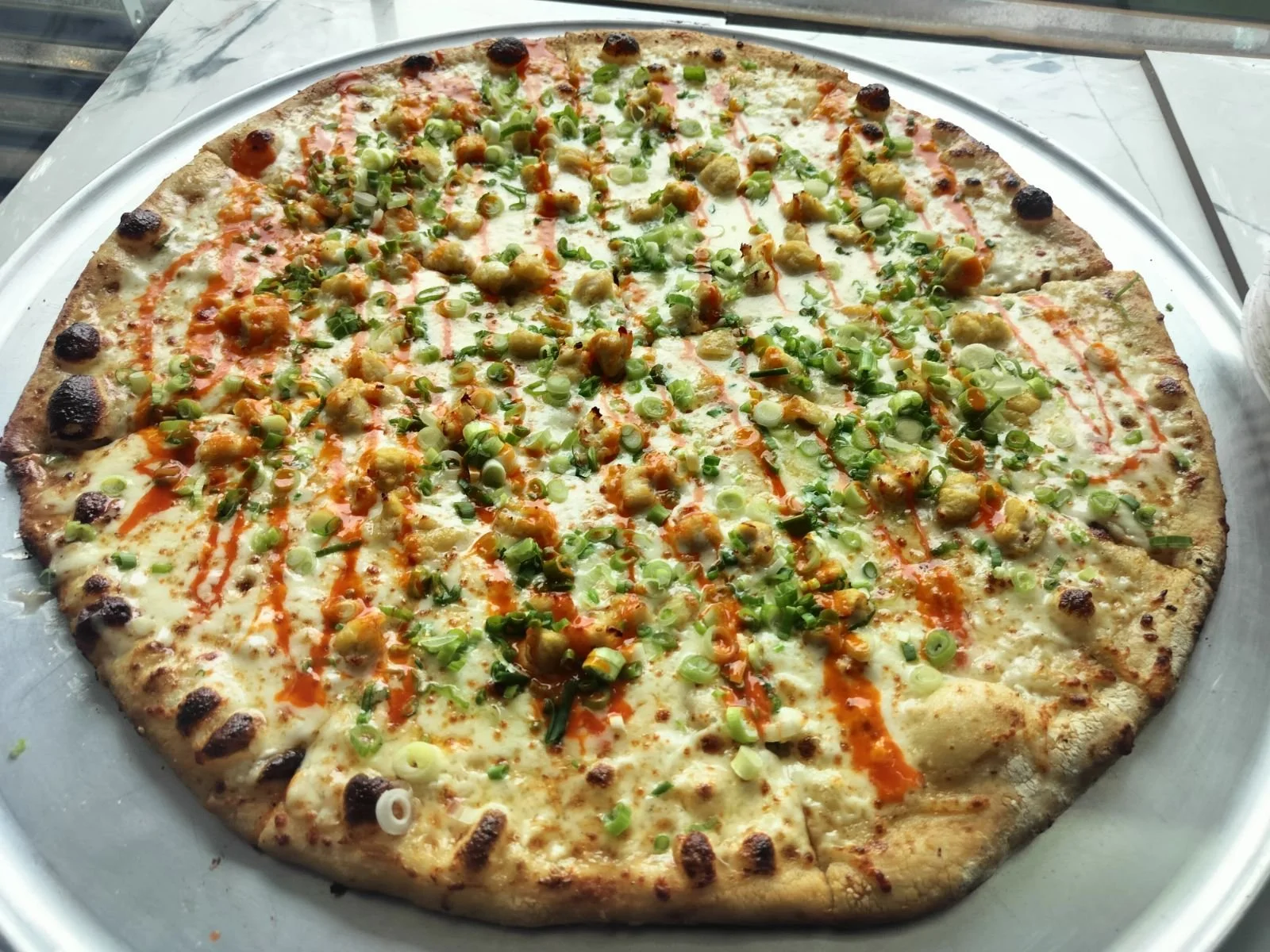 A large pizza with two halves, one topped with green onions, chicken, and hot sauce, and the other with mushrooms, cheese, and sauce, on a silver tray.