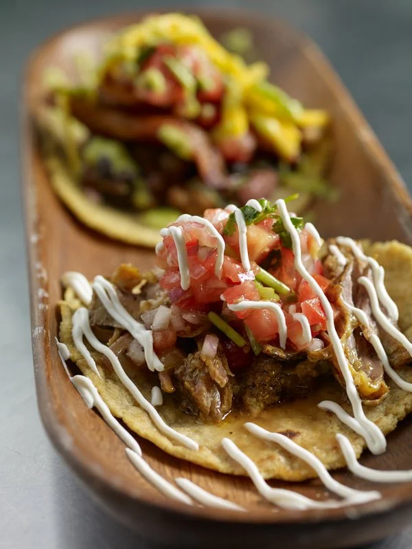 Two tacos on a wooden plate, one with shredded beef, vegetables, and white sauce, the other with shredded chicken, vegetables, and a yellow sauce.