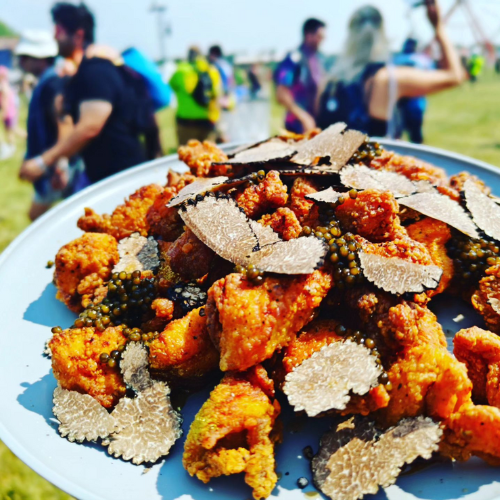 Fried chicken bites topped with black truffle slices on a plate at an outdoor event with people in the background.