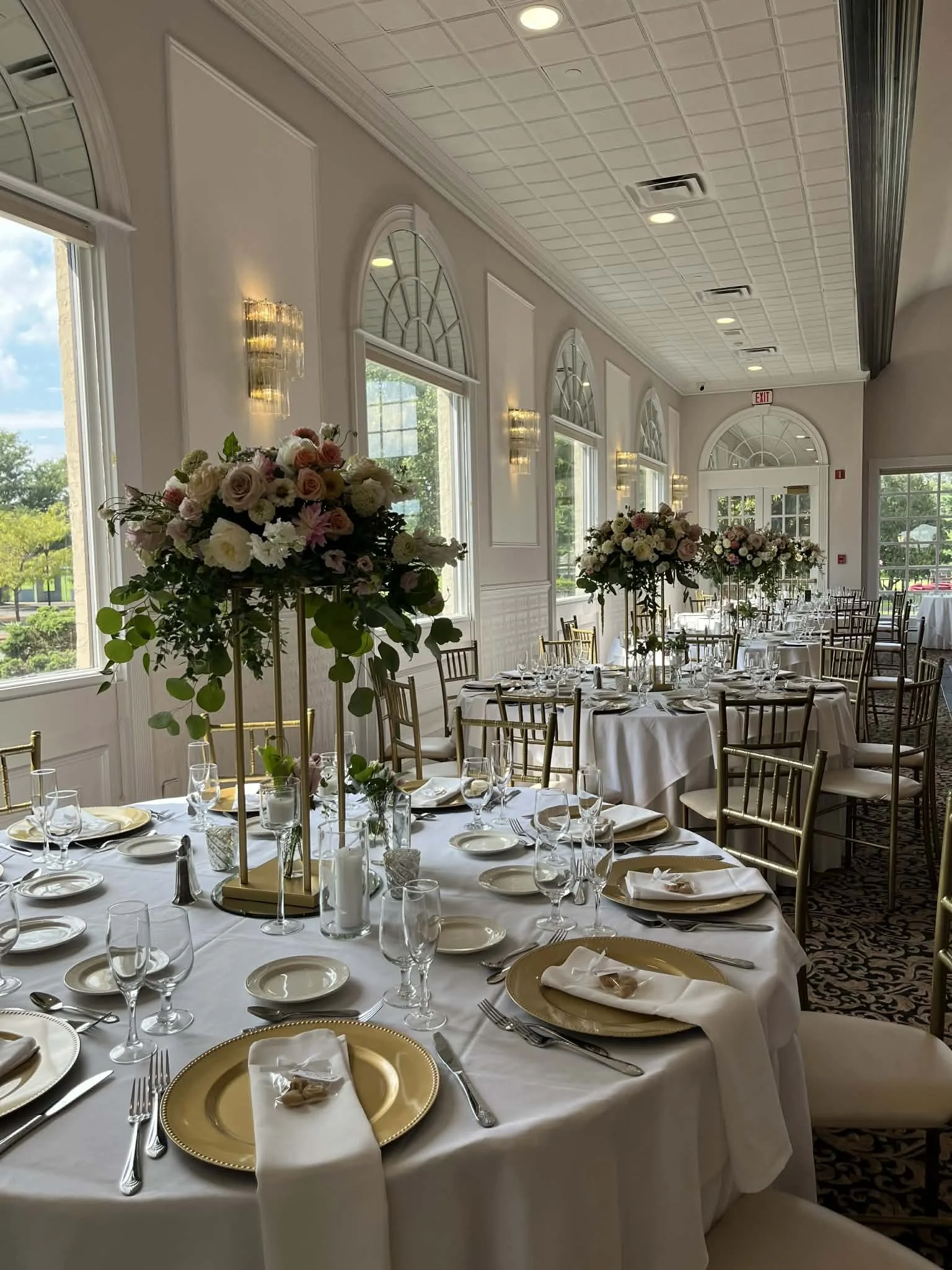 Elegant banquet hall with round tables draped in white tablecloths, decorated with tall floral centerpieces of roses and greenery, set with gold chargers, white napkins, and glassware, with large windows, arched mirrors, and soft lighting.