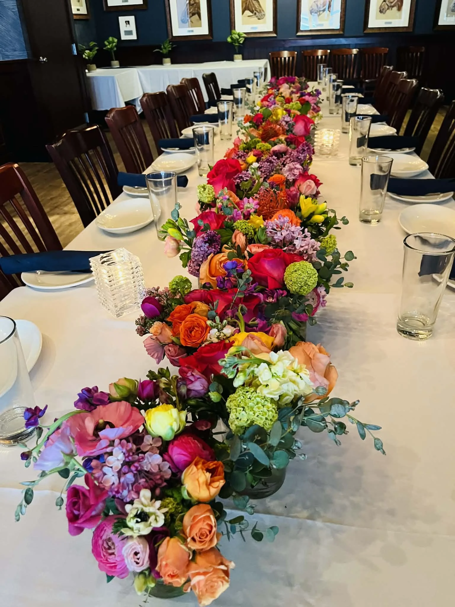 A long banquet table decorated with a vibrant, colorful floral centerpiece, set with white plates, blue napkins, clear water glasses, and surrounded by wooden chairs in a elegantly decorated room.