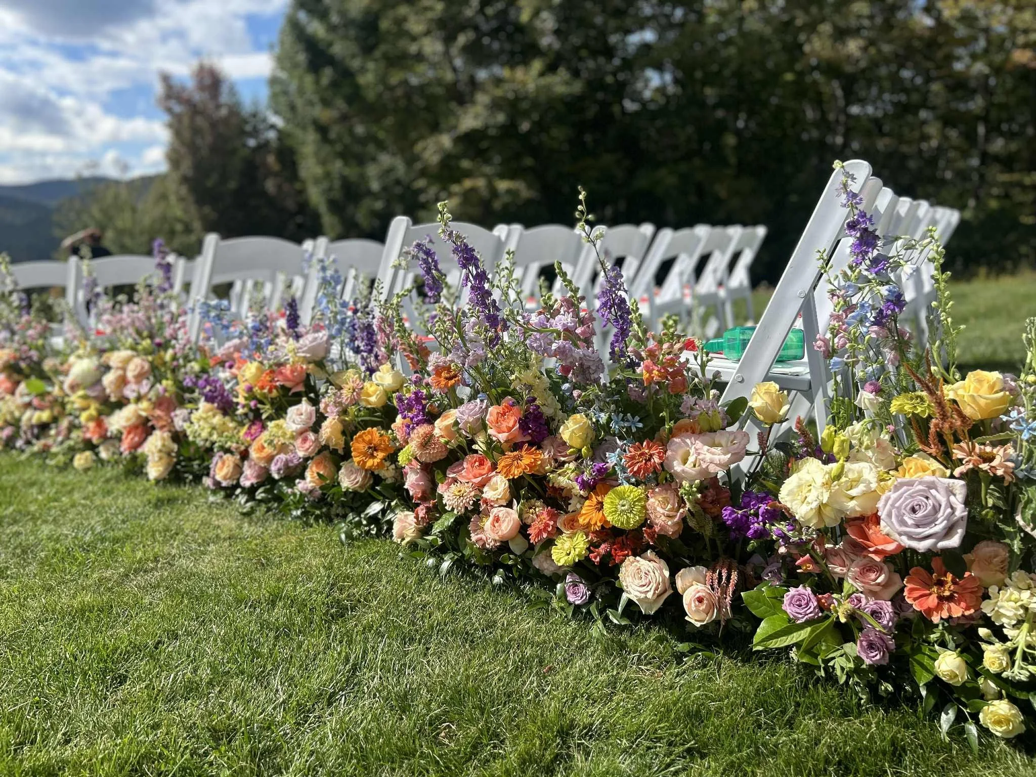 White chairs decorated with colorful flowers designed by Lot 32 Flower Farm on the grass outdoors, with trees and Adirondack mountains in the background.