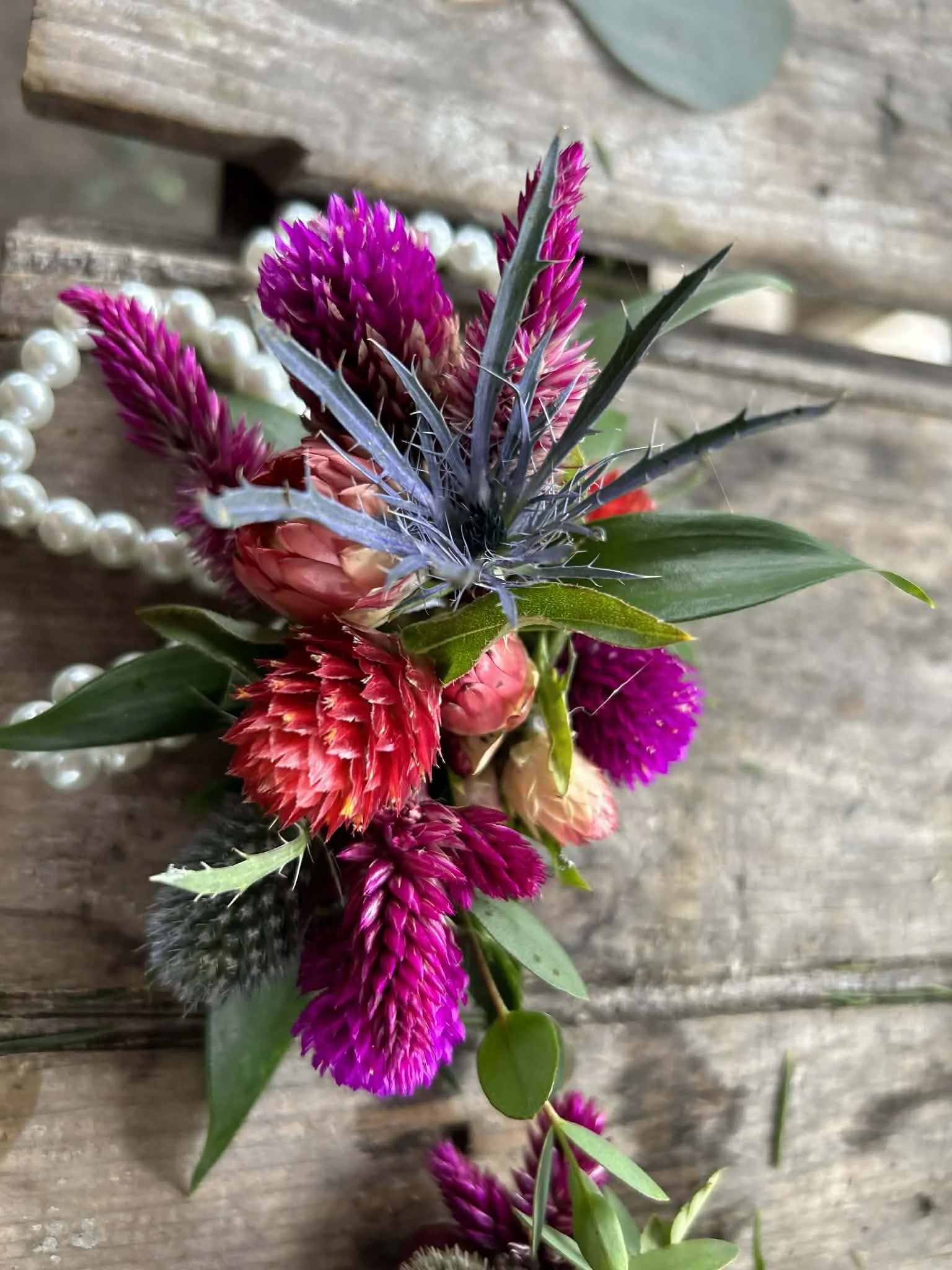 Close-up of a colorful bouquet of flowers with pink, purple, and red blooms, green leaves, and a pearl necklace, placed on rustic wooden surface.