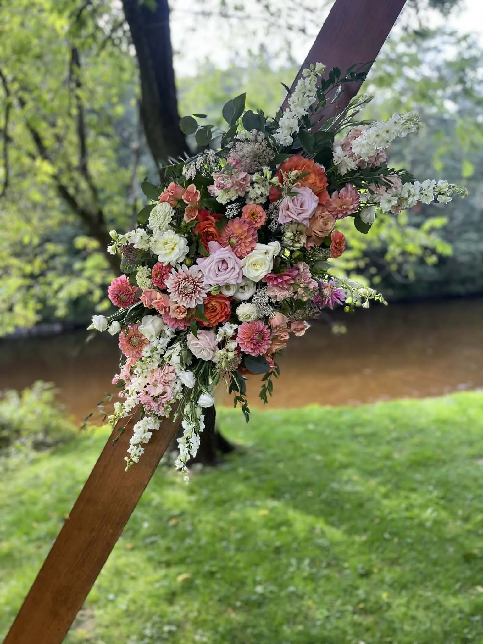 A large floral arrangement on a wedding arbor designed by Lot 32 Flower Farm with pink, white, and orange flowers attached to a wooden post outdoors near a river, with trees and grass in the background.