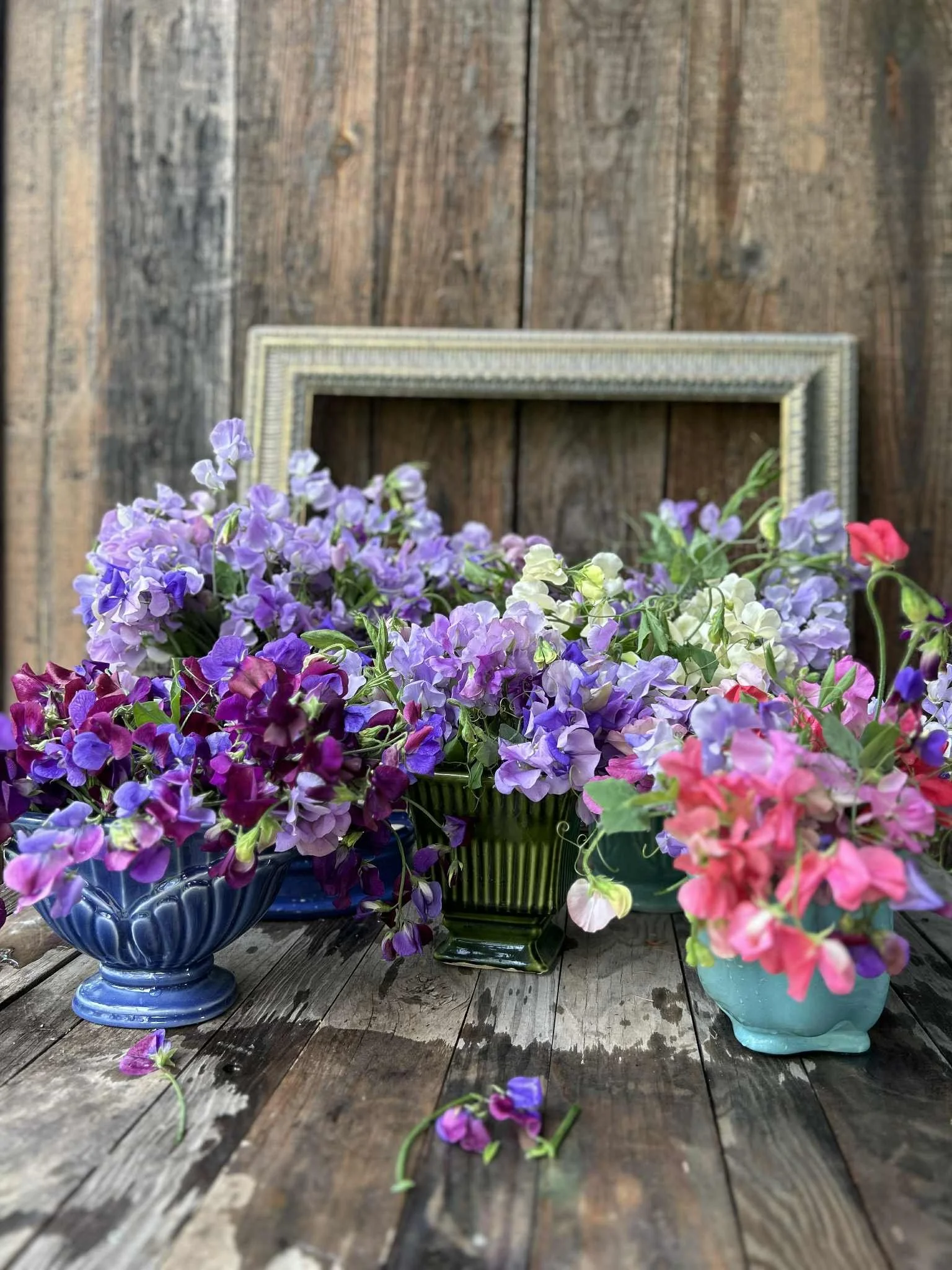 Arrangement of purple, pink, and white flowers in ceramic, glass, and metal vases on a rustic wooden table behind a wooden wall with an empty picture frame.